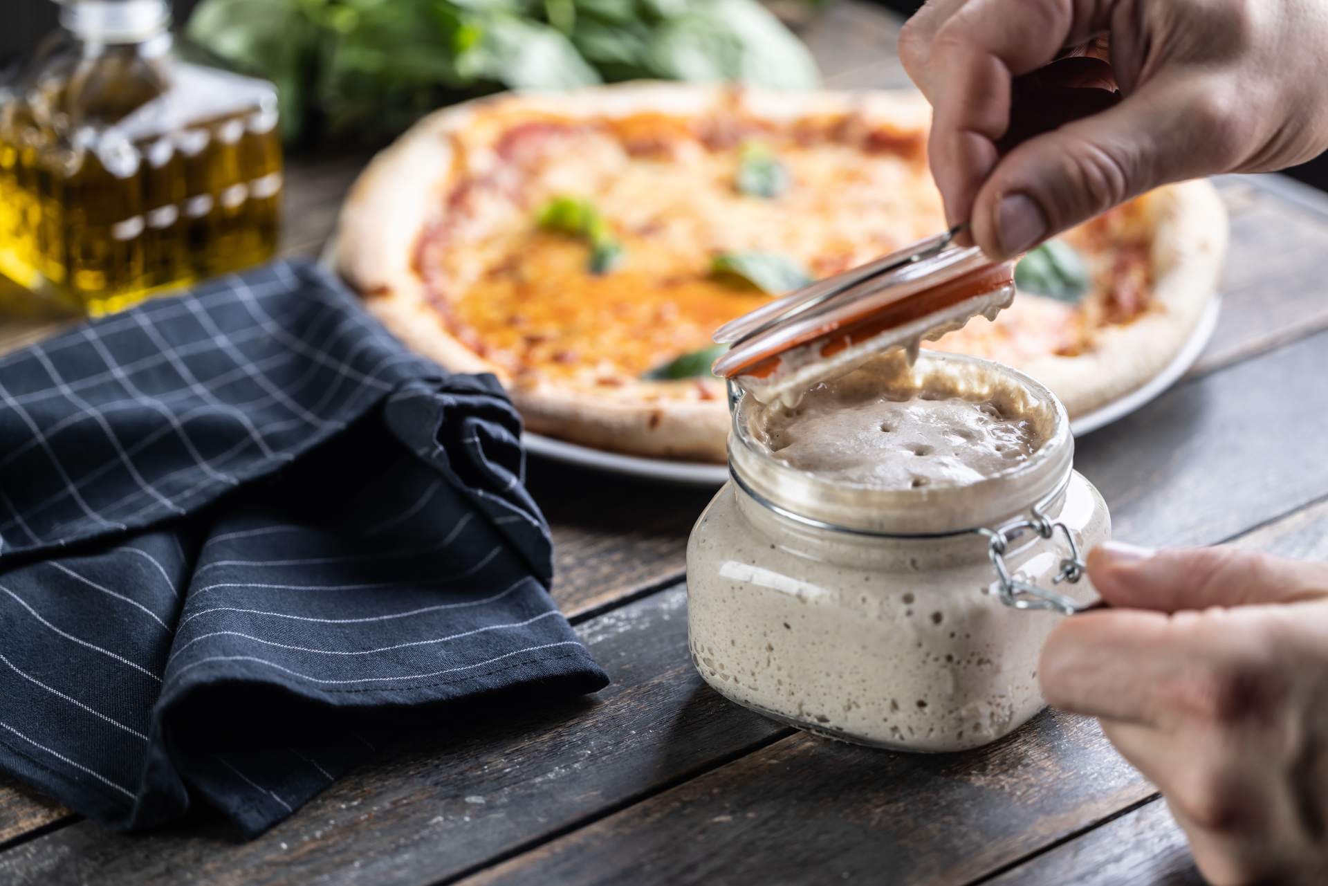 Sourdough starter in a glass jar on a table with a cooked cheese pizza with basil in the background.