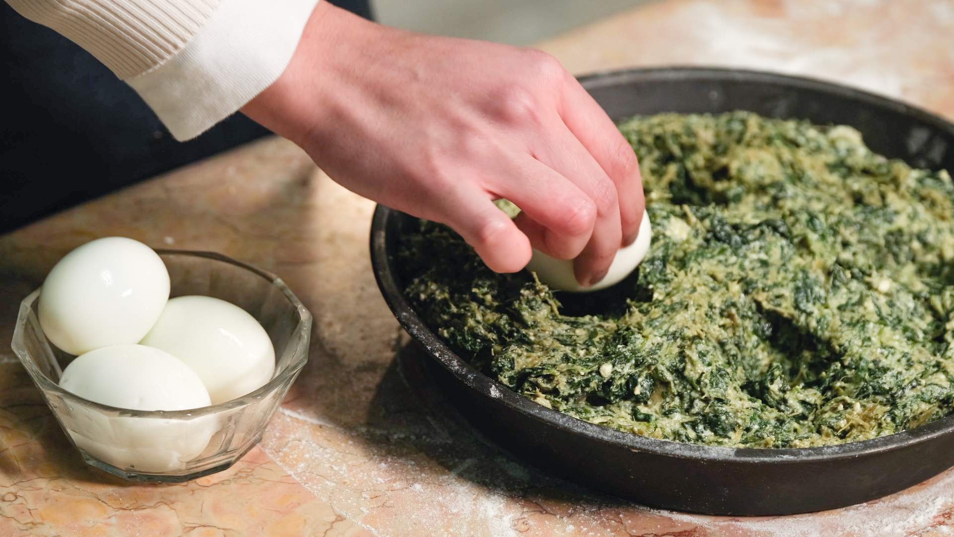 Boiled egg being placed into a mixture of spinach and cheese in a cake tin filled with pastry