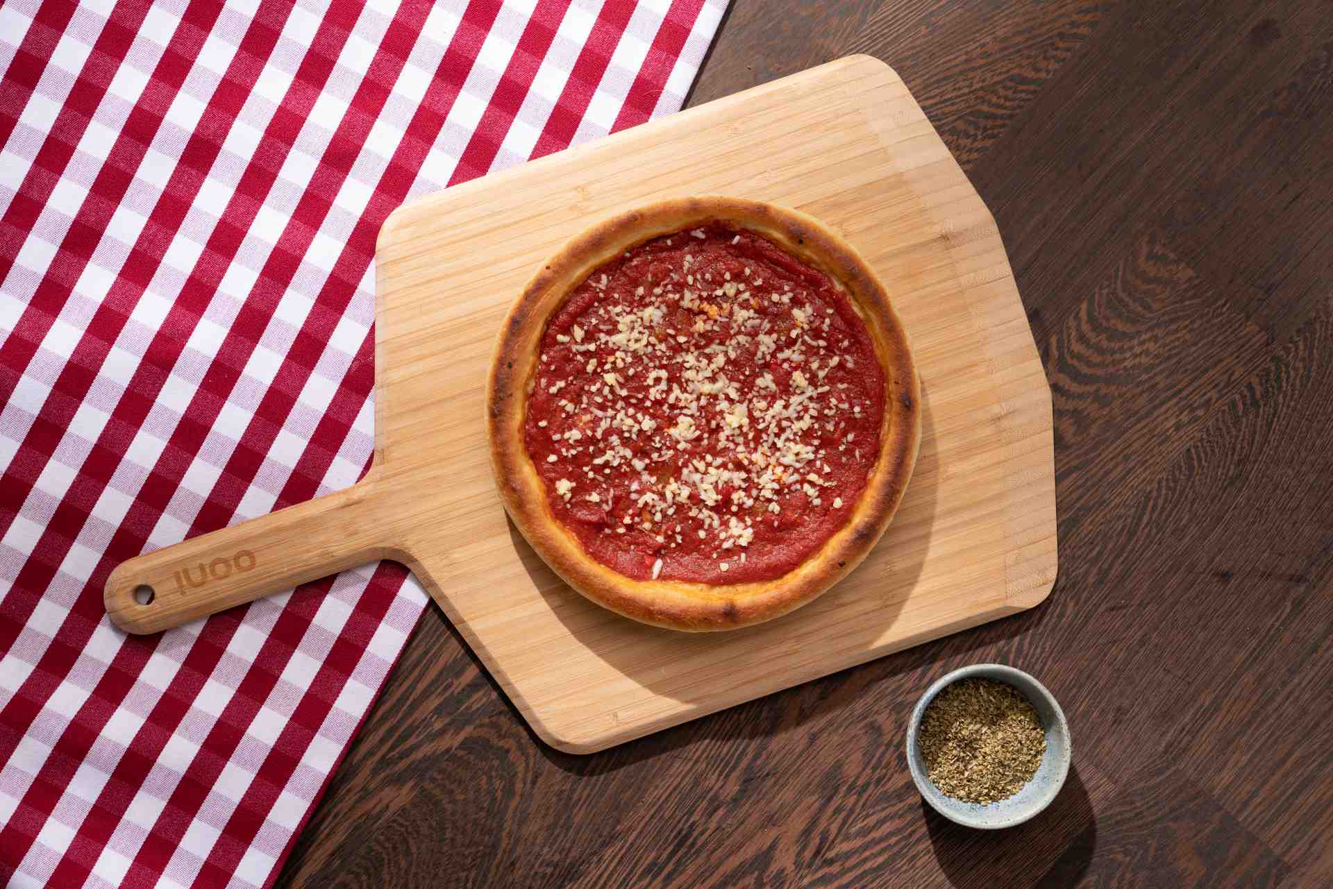 Chicago deep-dish pizza with tomato sauce and shredded mozzarella on an Ooni Bamboo Pizza Peel & Serving Board next to a bowl of oregano.