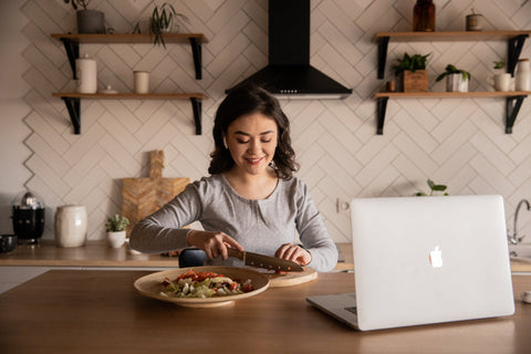 Woman cutting vegetables in her kitchen with an open laptop