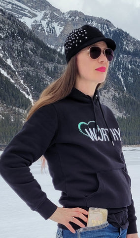 Woman wearing a black hoodie with 'Worthy' on the front & a black hat in front of mountains