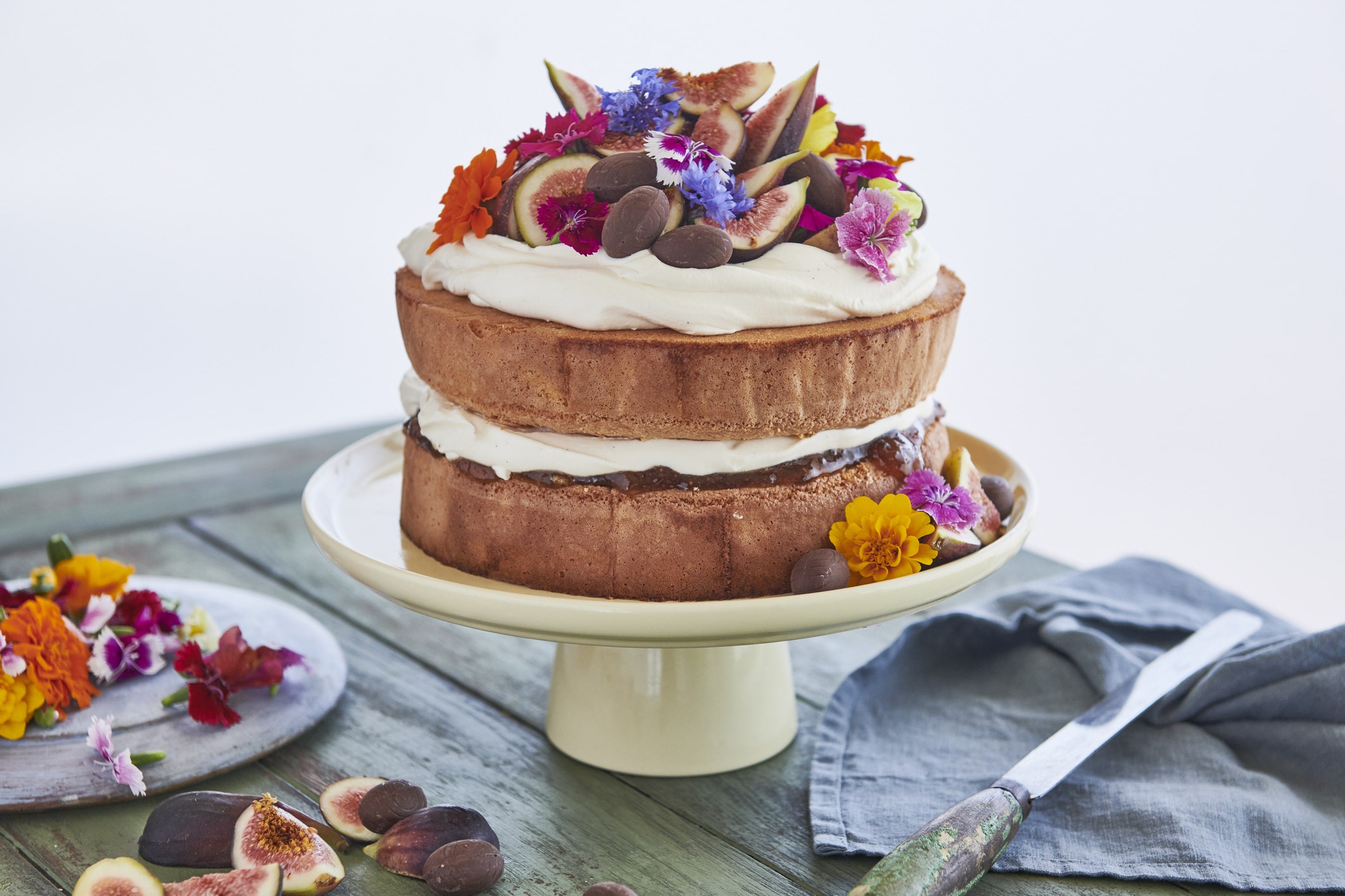 Traditional Italian Easter Sponge Cake on a white cake stand.  Decorated with fresh figs, edible flowers and dark chocolate eggs.  A plate of fresh flowers to the left of the stand and fresh quartered figs in the foreground.