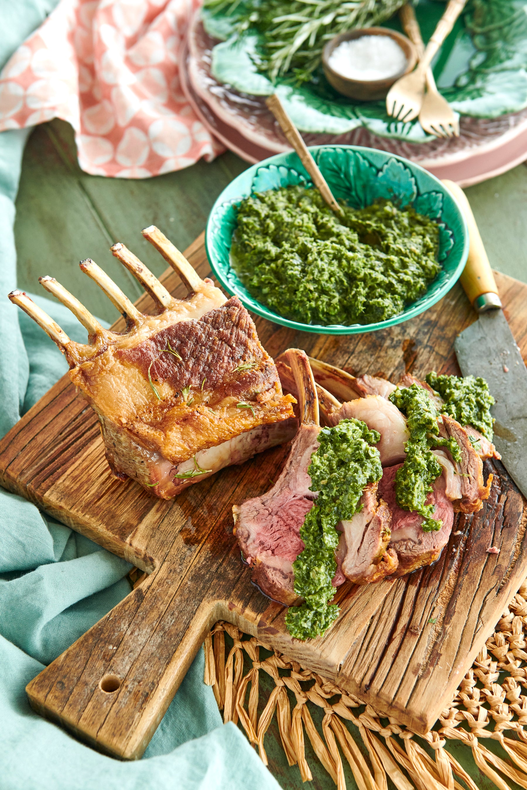 Blue bowl at top right that contains salsa verde.  Cooked rack of lamb underneath with some lamb cutlets on a wooden chopping board and covered with salsa verde.
