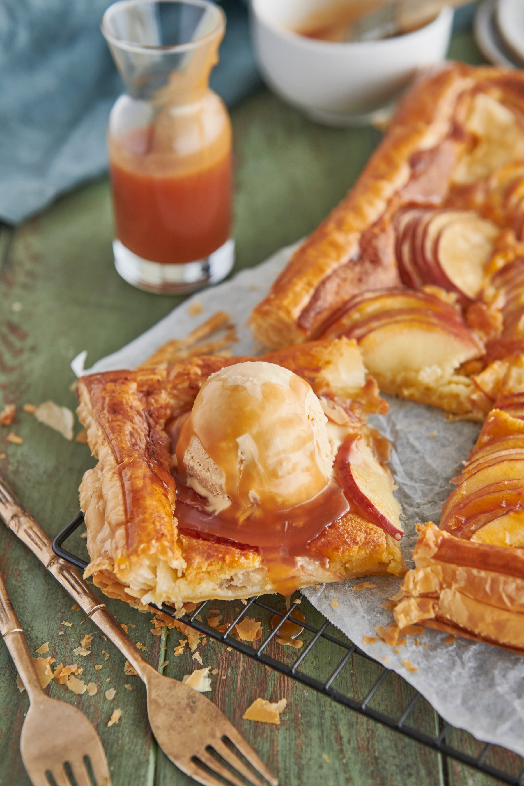 Cooked apple galette with a slice cut out of it.  The slice has a scoop of ice cream and a drizzle of caramel sauce on it. 2 golden forks as in the bottom left corner.