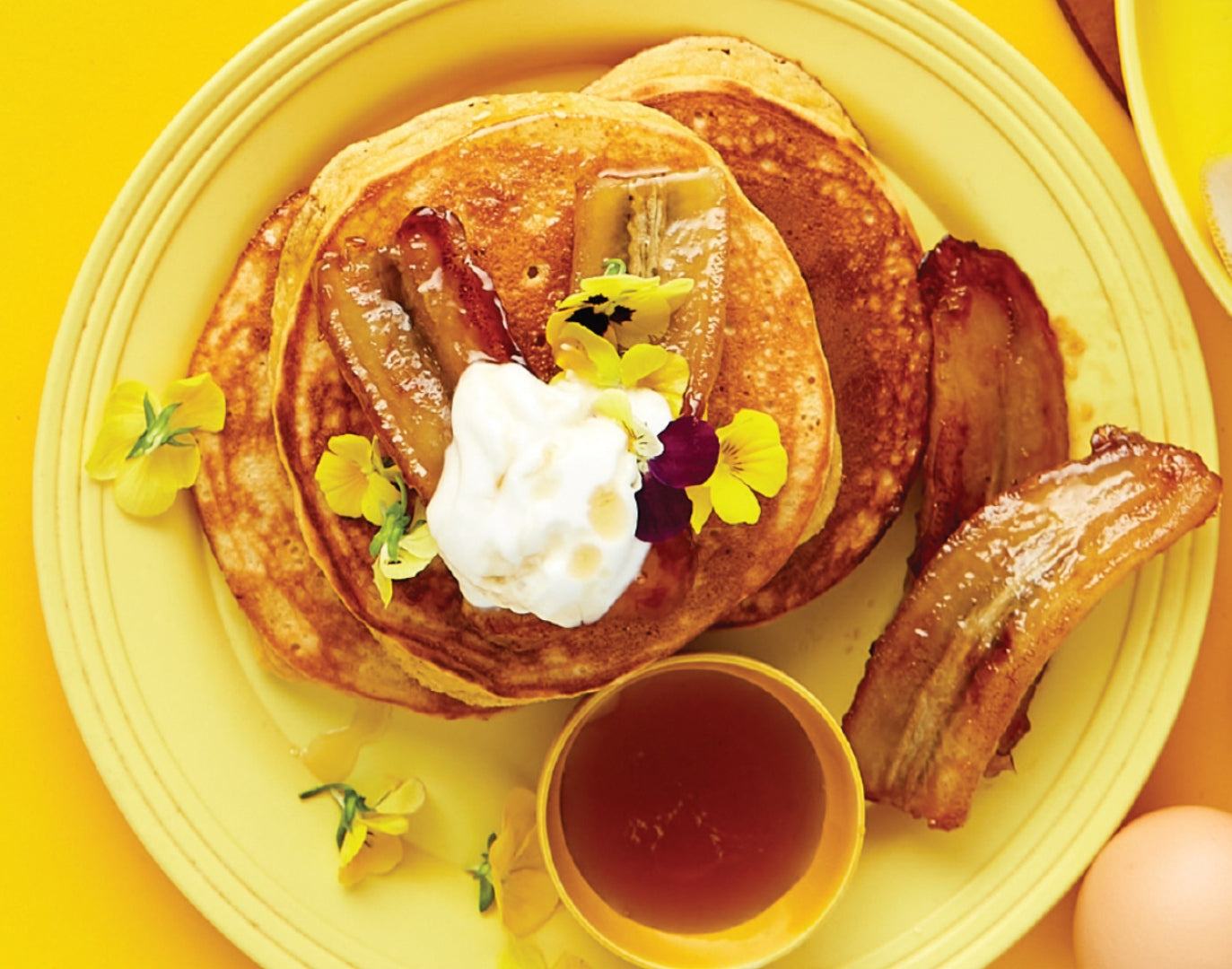 A yellow plate with pancakes, yoghurt, maple syrup and caramelised bananas.