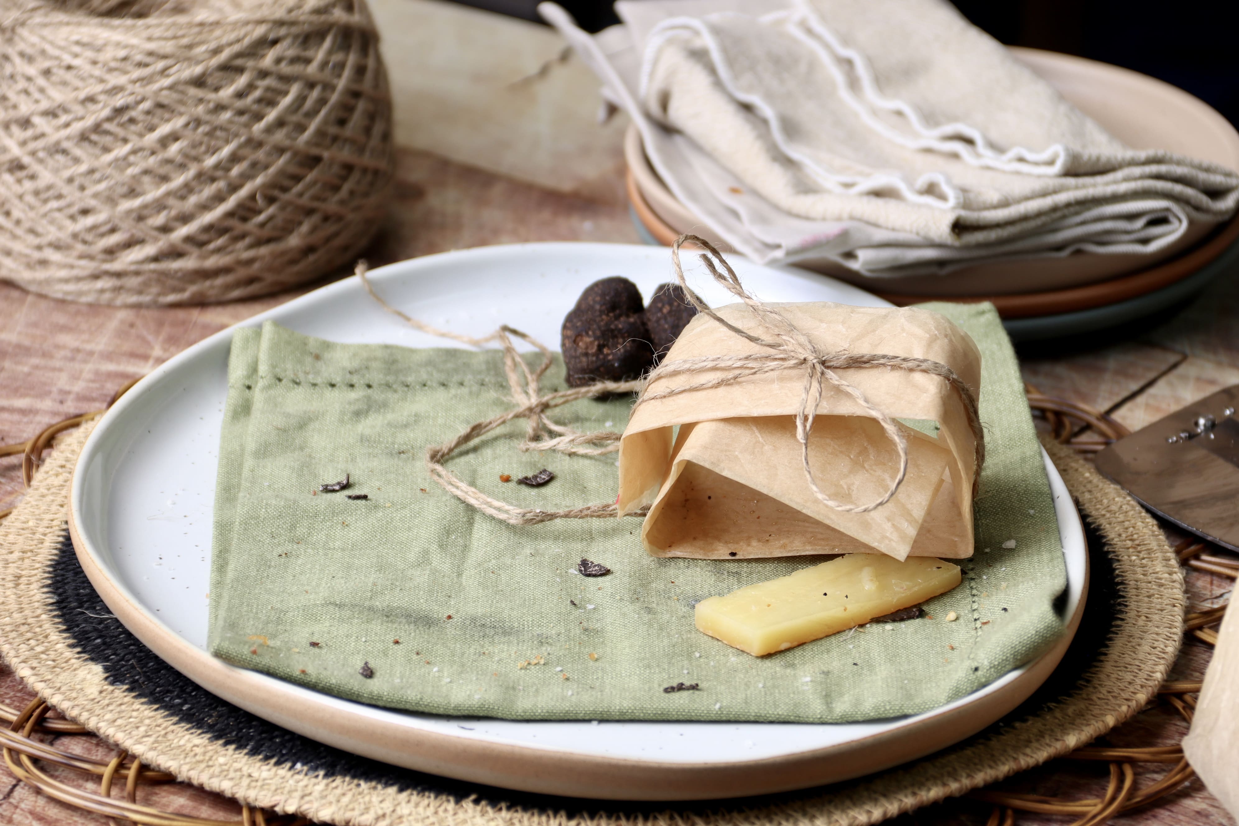 Empty truffle toastie plate with remaining baking paper tied with kitchen twine.  Half a fresh black truffle in the background and a cheese rind in the foreground.