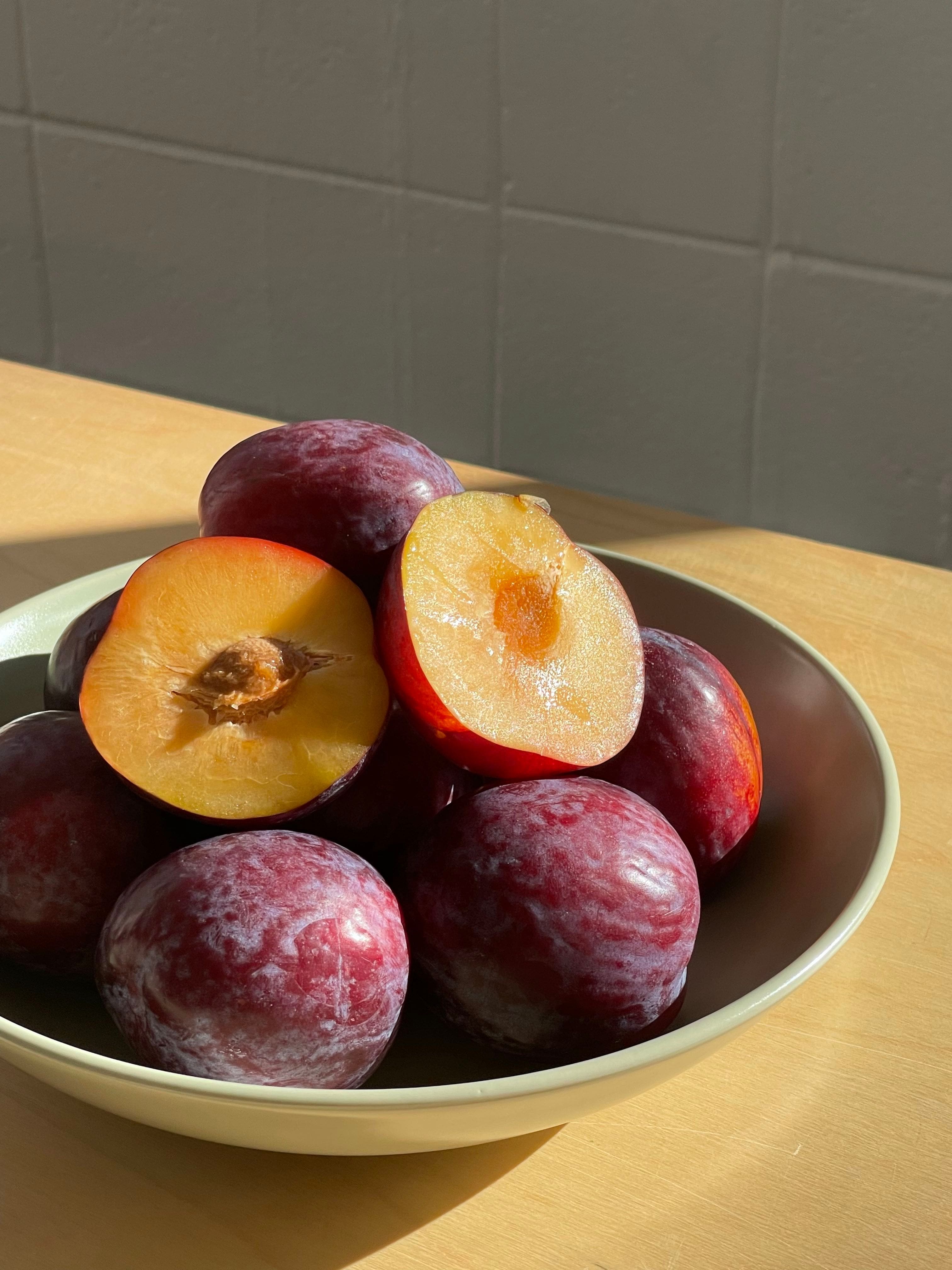 Bowl of flavour fall plums on a wooden bench with one cut in half sitting on top.