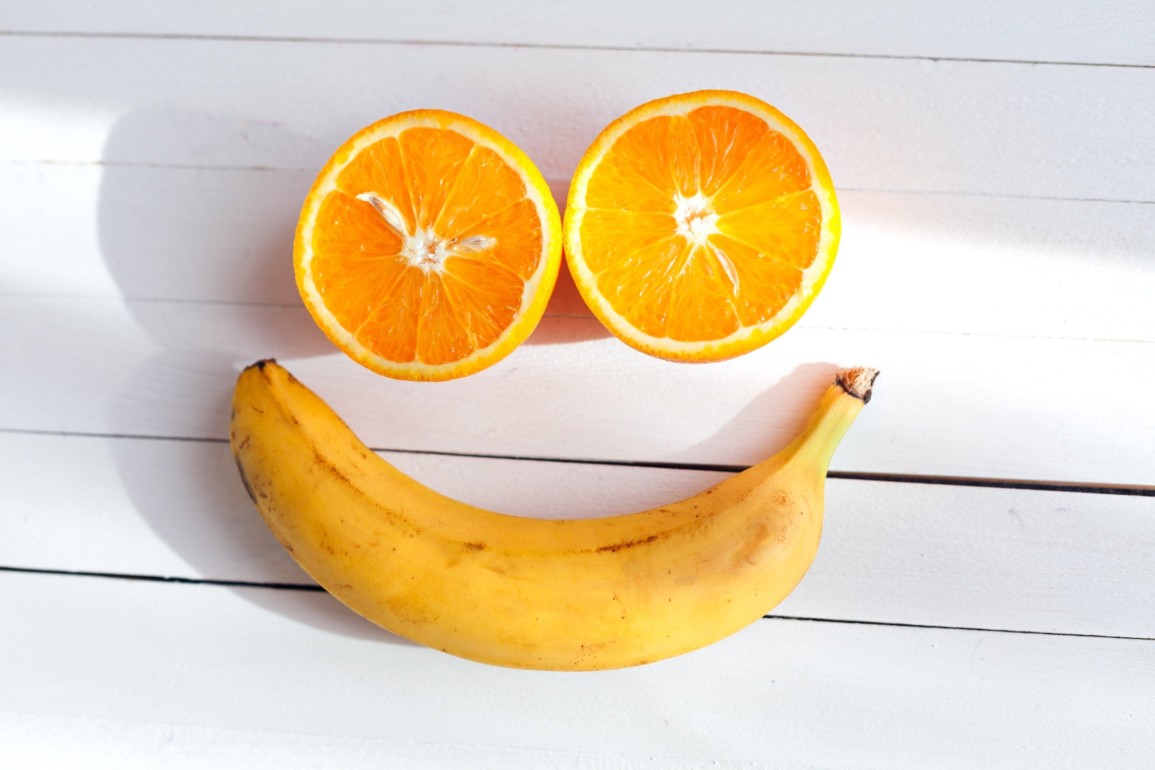 A smily face made from a banana and halved orange against a white wooden table