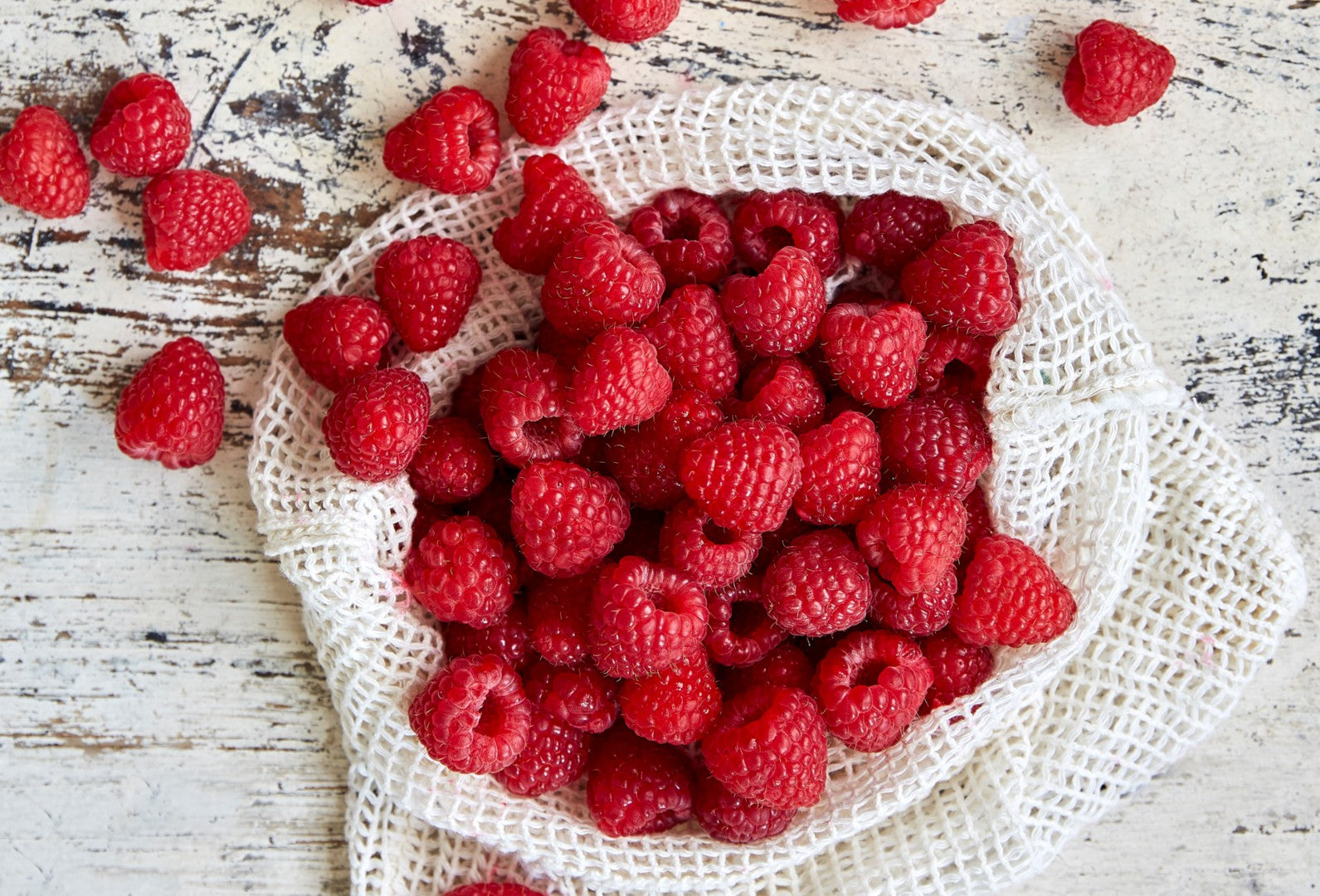 A white net bag of raspberries on a worn white wooden table