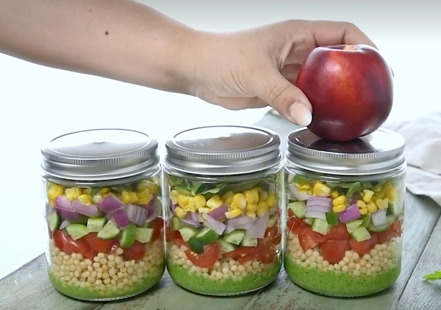 A picture of three jars full of layered salad with a woman's hand adding a nectarine to the top of one jar