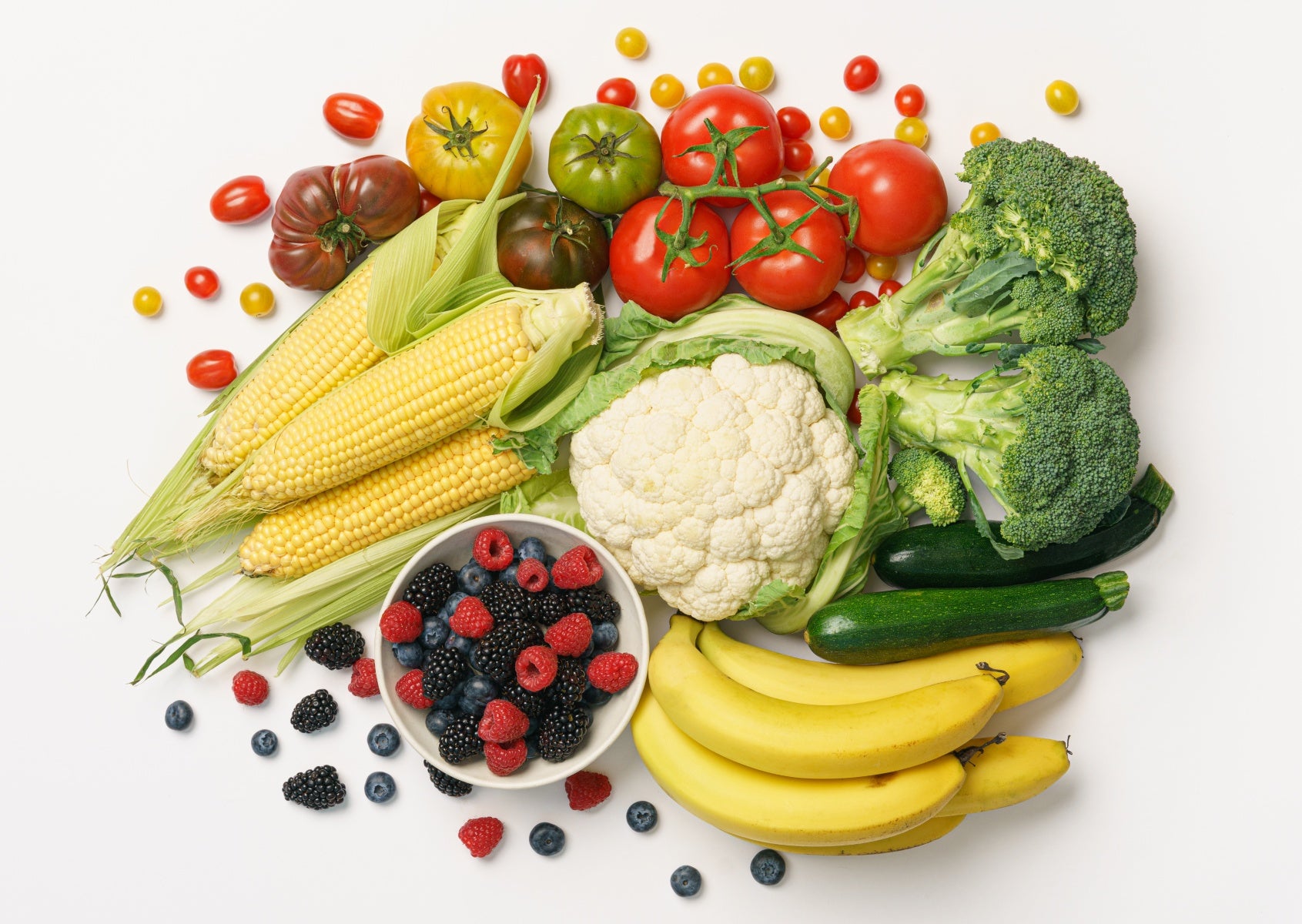 A photograph of bananas, broccoli, berries, cauliflower, corn, tomatoes and zucchini on a white background