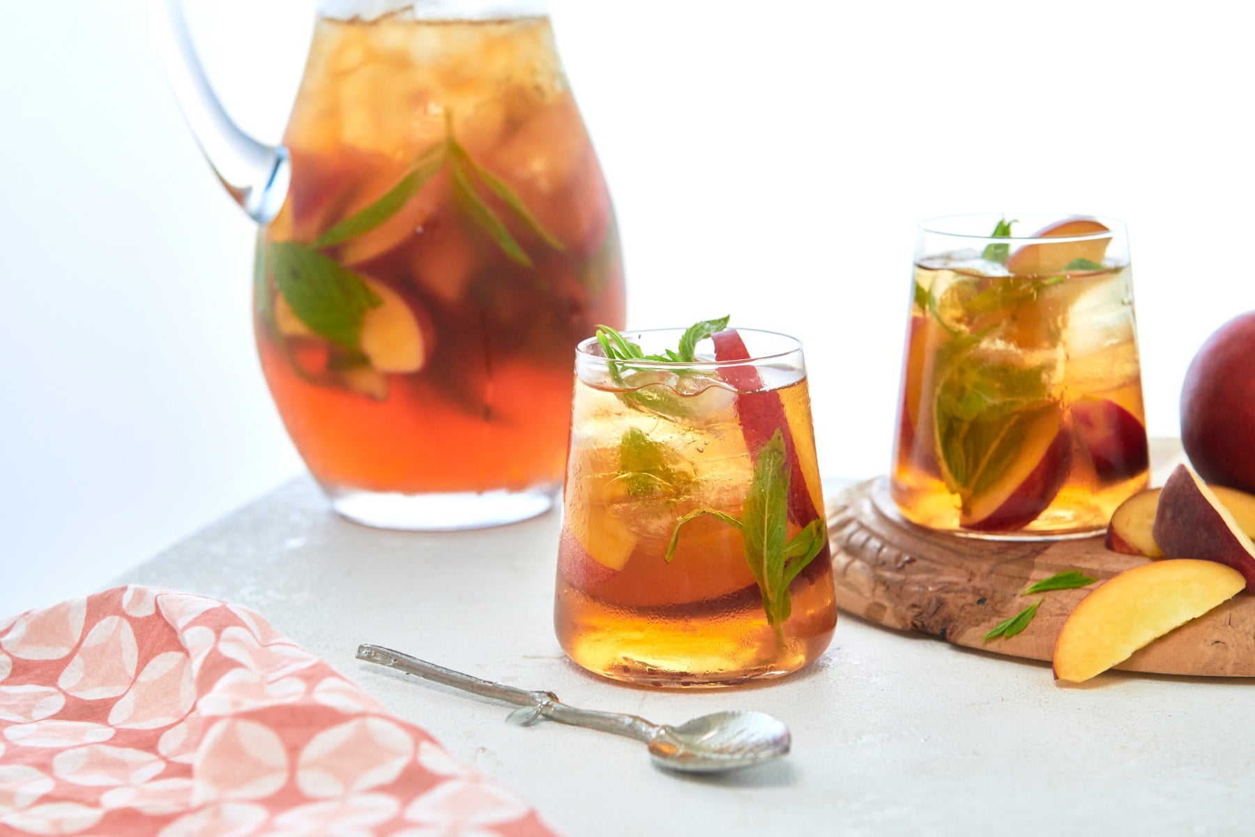 A jug and two glasses filled with homemade peachy iced tea, including slices of stone fruit and mint leaves