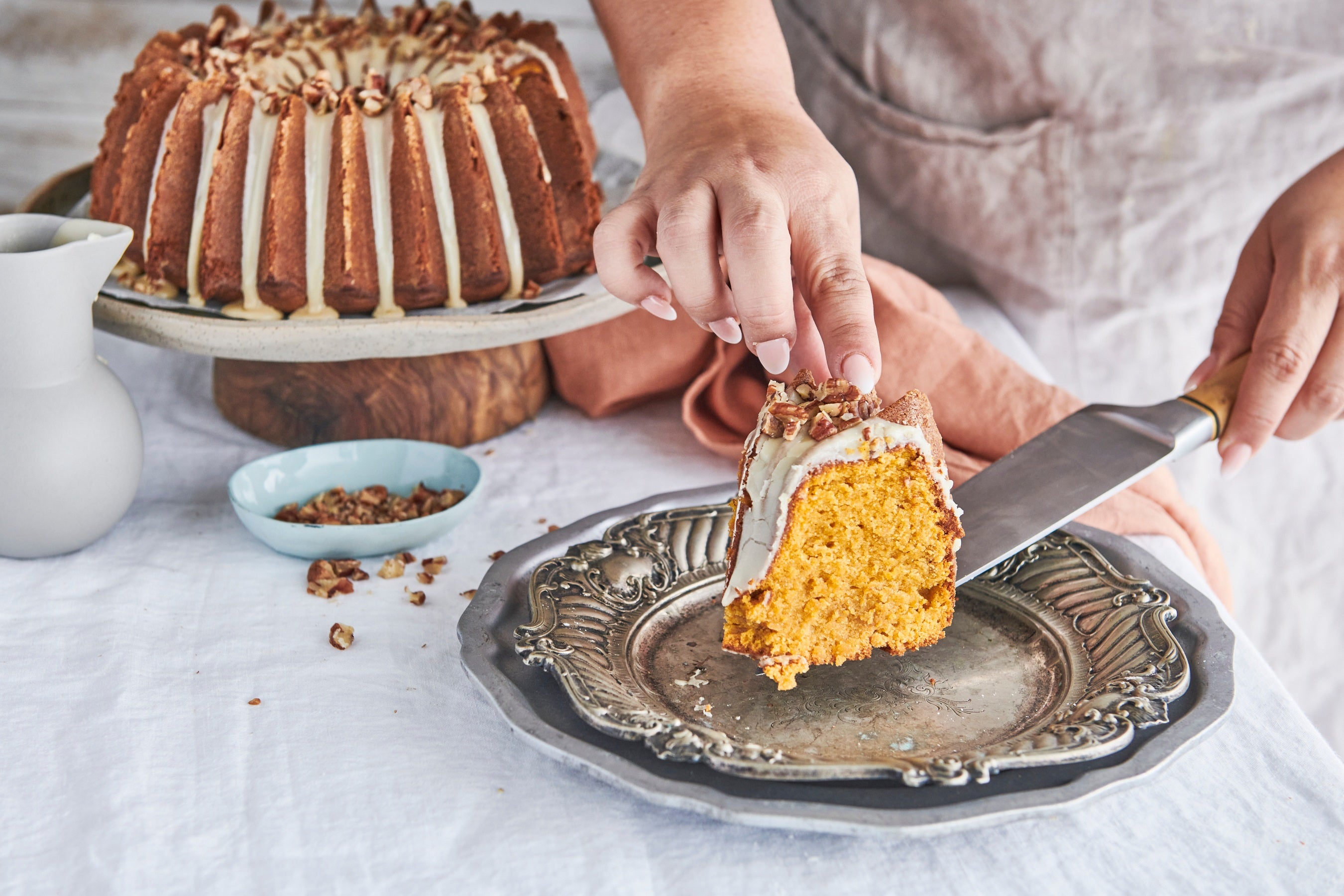 A woman is serving a slice of pumpkin spice bundt onto a silver plate, the bundt is in the background covered with maple syrup glaze.