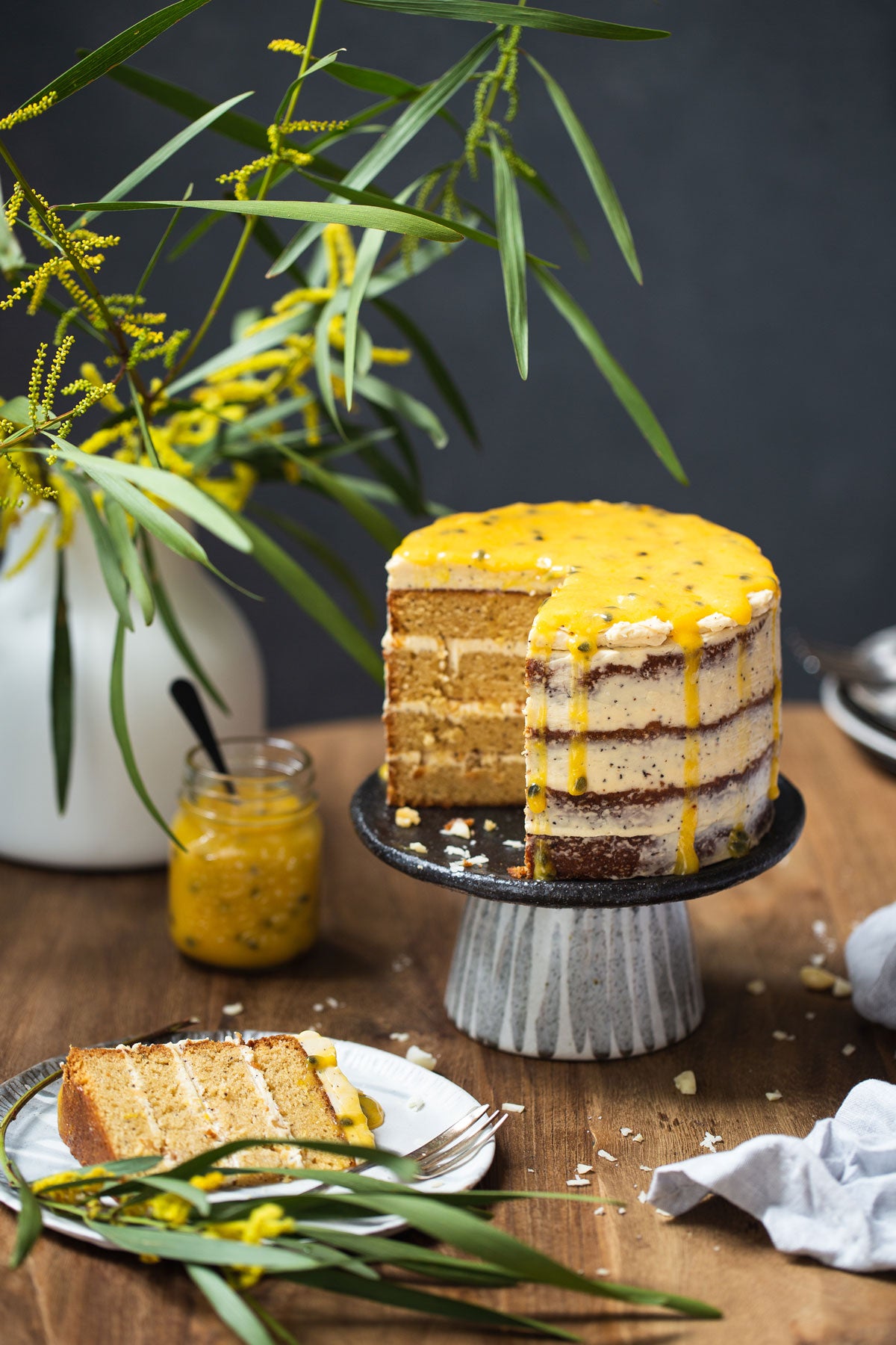 The cake is in the background and a slice of the cake is on the plate in front.  There is a mason jar of passionfruit curd in the background and behind that is a bunch of wattle.