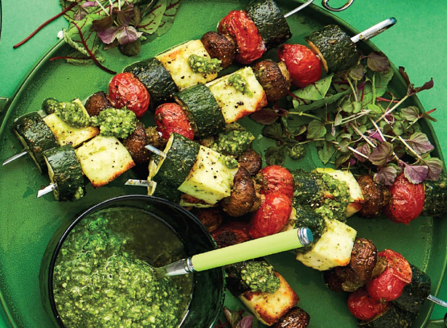 A green plate piled with zucchini and halloumi skewers with a small bowl of basil pesto