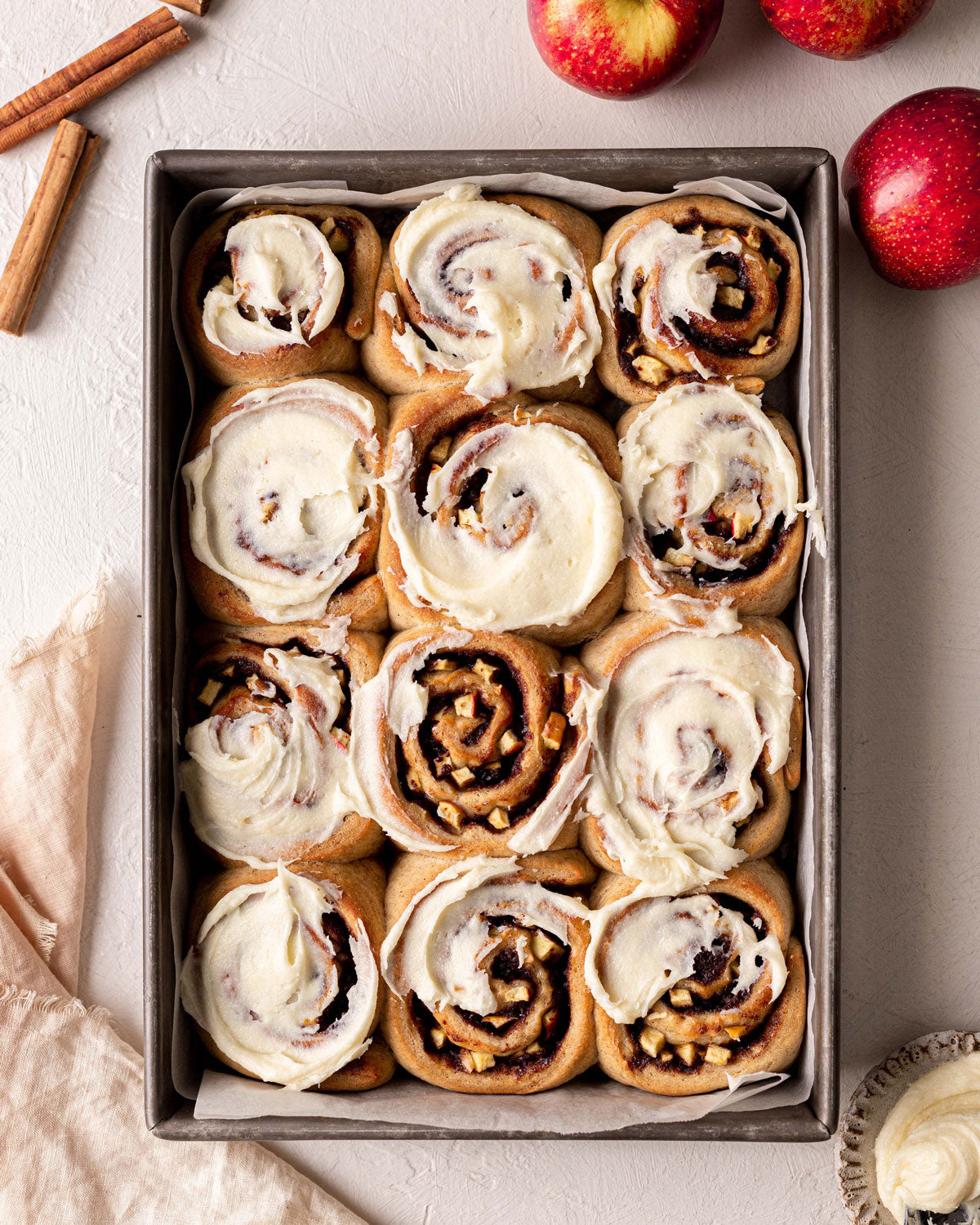 Vegan Apple Cinnamon Scrolls with vegan cream cheese icing.   Photo shows 12 scrolls in a baking pan surrounded by red apples and cinnamon quills