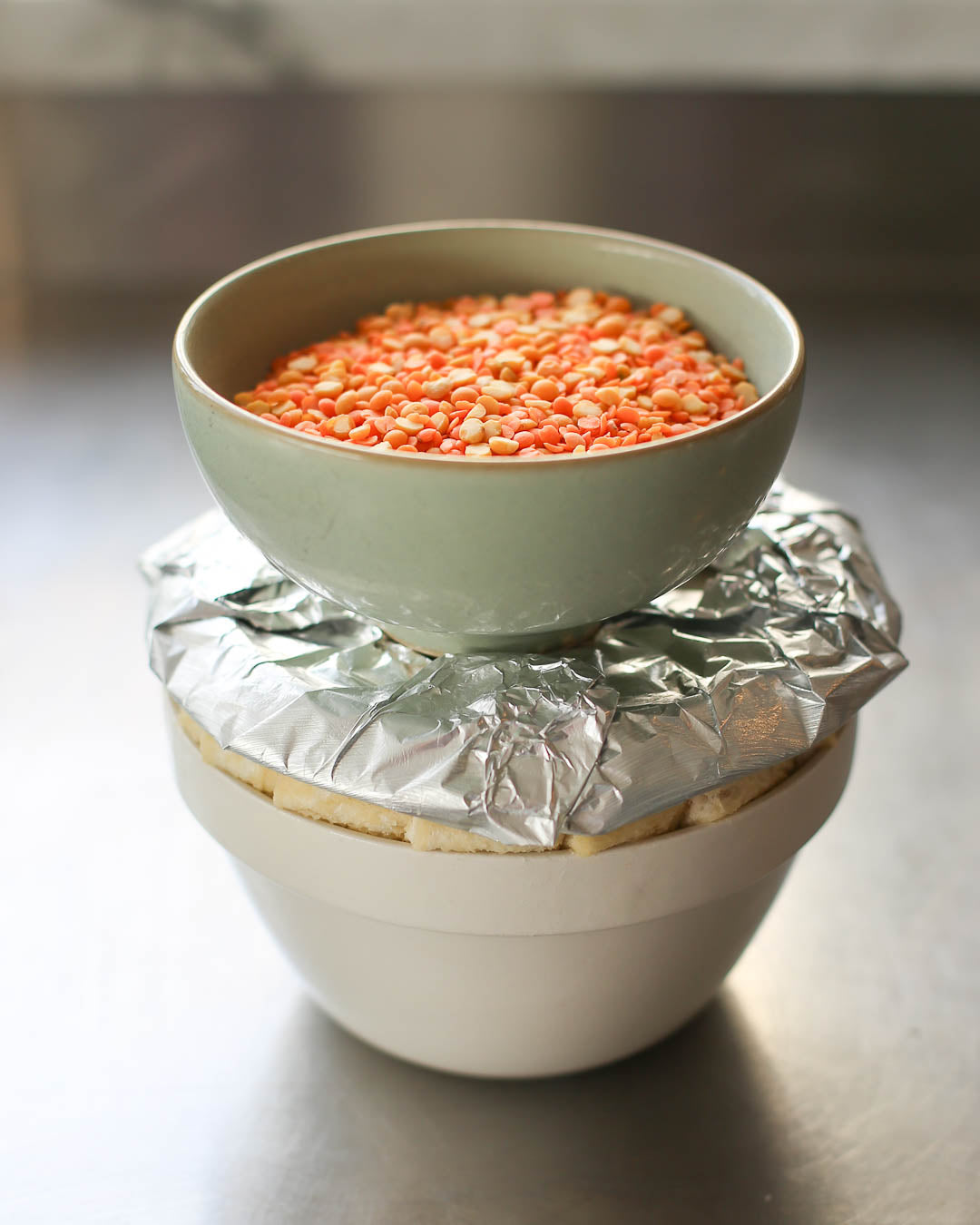 Image shows the filled bread basin, covered with foil and a bowl of red lentils acting a a pastry weight on top.  This is how it should go into the oven to cook.