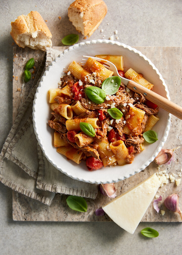 White bowl of lamb shoulder ragu with rigatoni.  Garnished with 4 basil leaves.  There is a wedge of cheese and 3 garlic cloves in the bottom right corner and two pieces of baguette with 2 basil leaves in the top right corner.