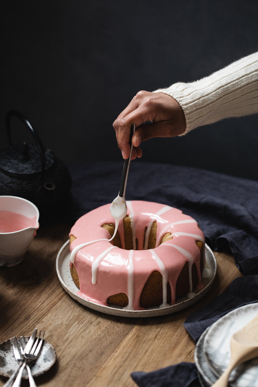 Blood Orange and Earl Grey Bundt Cake with pink and white striped icing.  There is a hand and arm in the top right corner holding a spoon that is dripping with white icing.