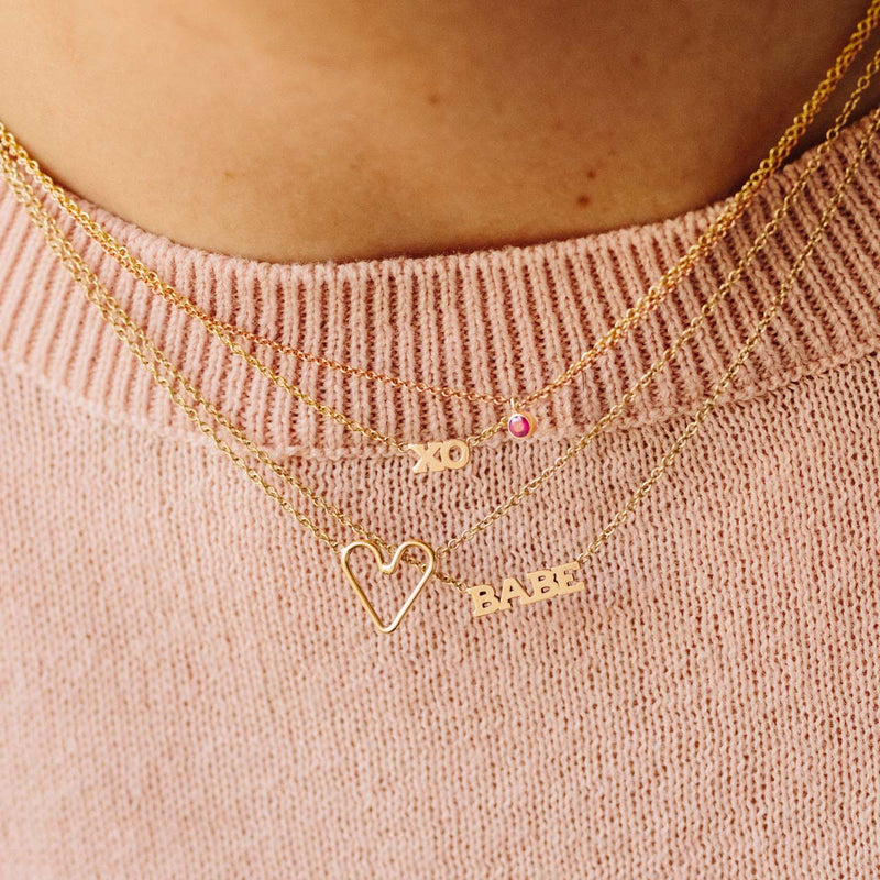 woman in pink sweater wearing Zoë Chicco 14kt Gold Itty Bitty BABE Necklace, Itty Bitty XO Necklace, Tiny Open Heart Necklace, & Ruby Pendant Necklace layered together