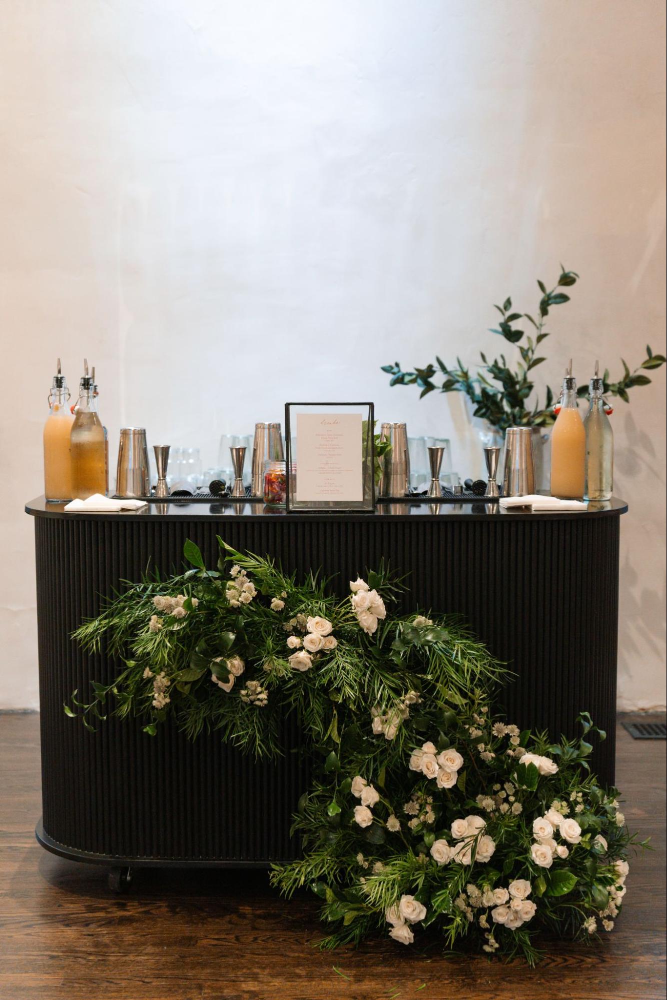 Serving a custom cocktail, a black bar stand is decorated with fresh, white and green florals.