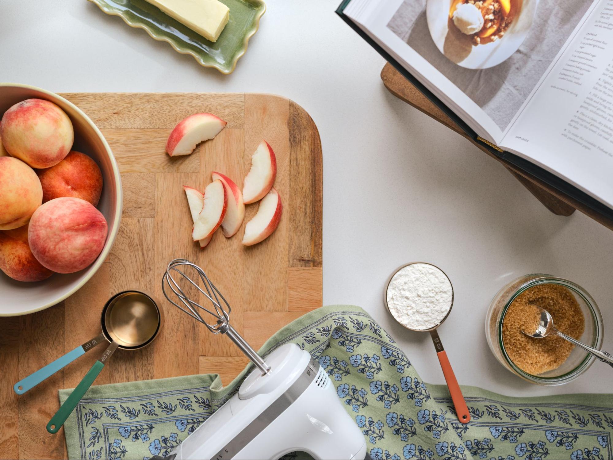 Peaches, flour, and sugar are laid out with Magnolia Table Cookbook open to a recipe for Peach Cobbler.