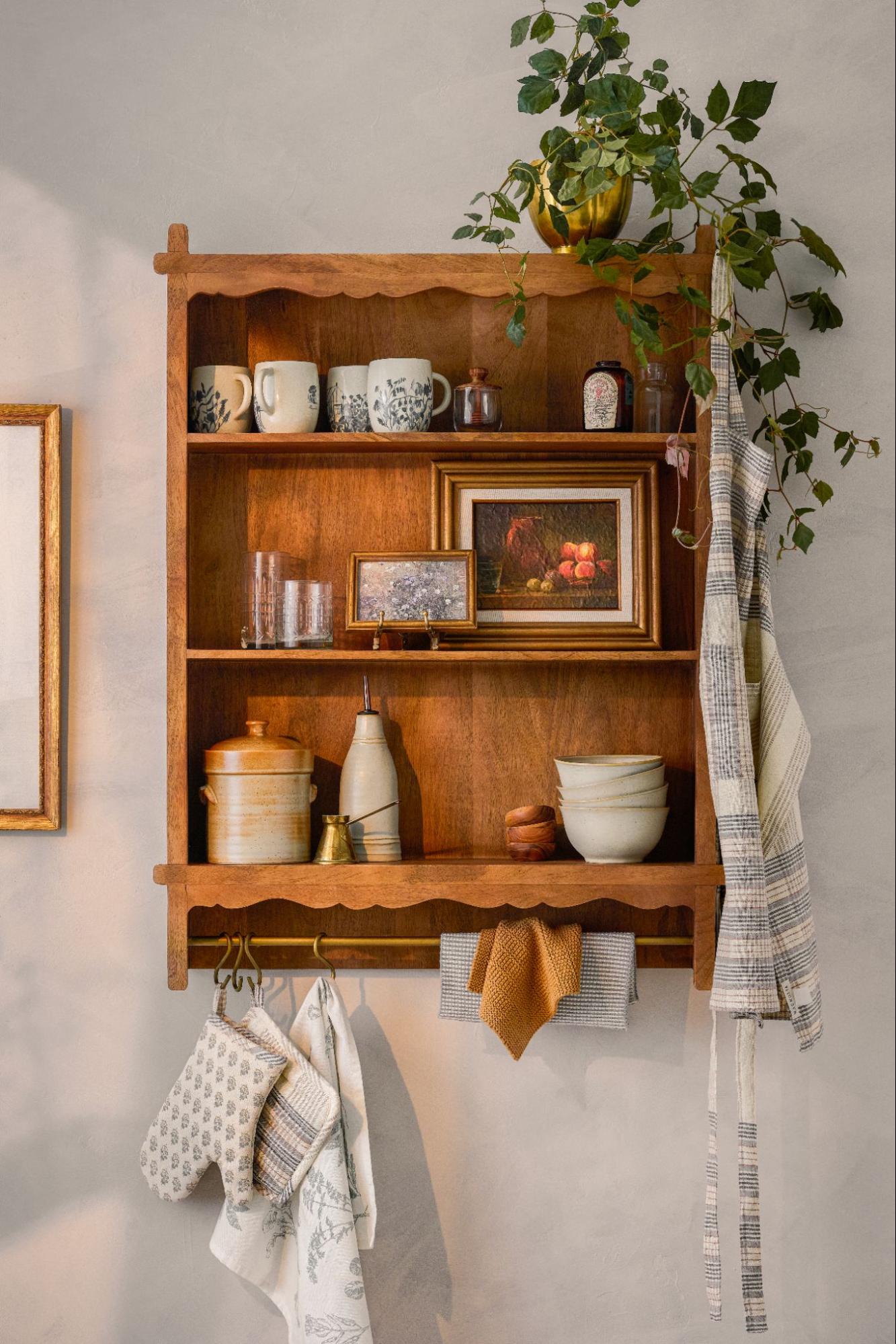 A wooden wall shelf is filled with kitchen pieces and decor. A hook rail is lined with kitchen linens and pot holders.