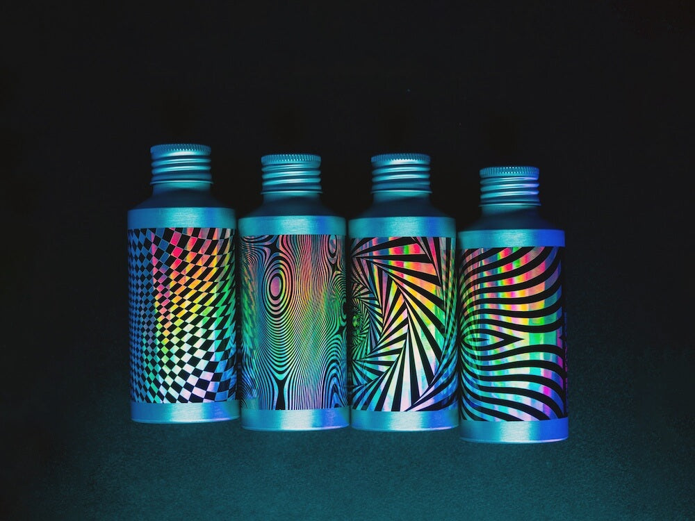 Four aluminum Beardbrand Utility Oil bottles in a untidy row on a dark surface, with holographic labels and black psychedelic designs in dim lighting.