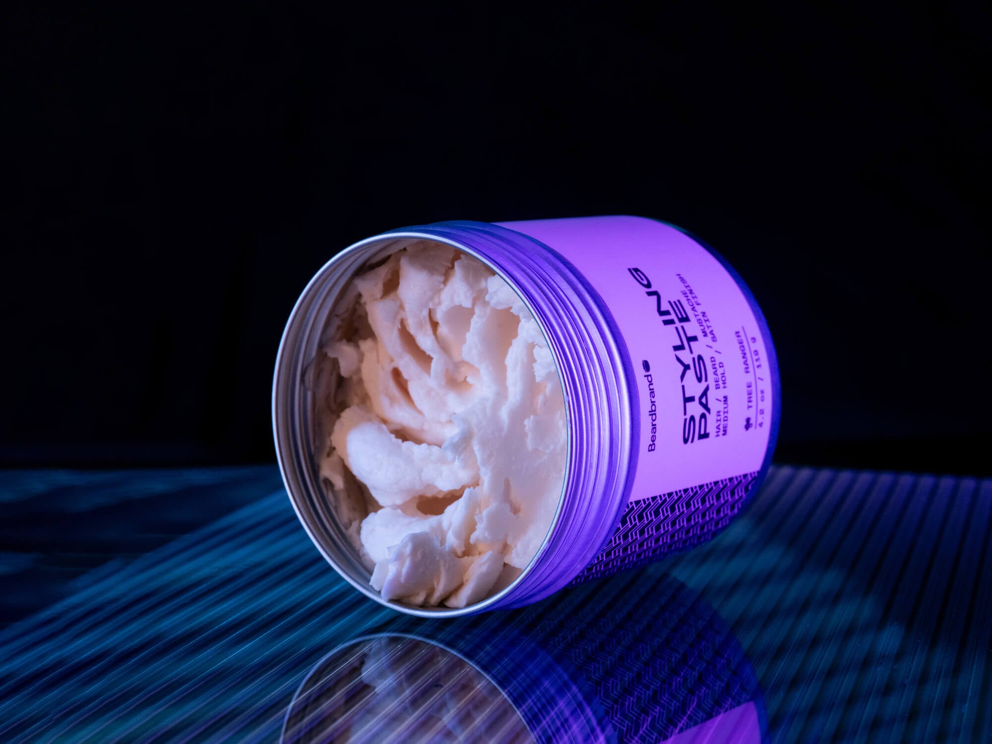 A container of Beardbrand Styling Paste under purple lighting, lying on its side with the lid off, revealing the creamy white product inside.