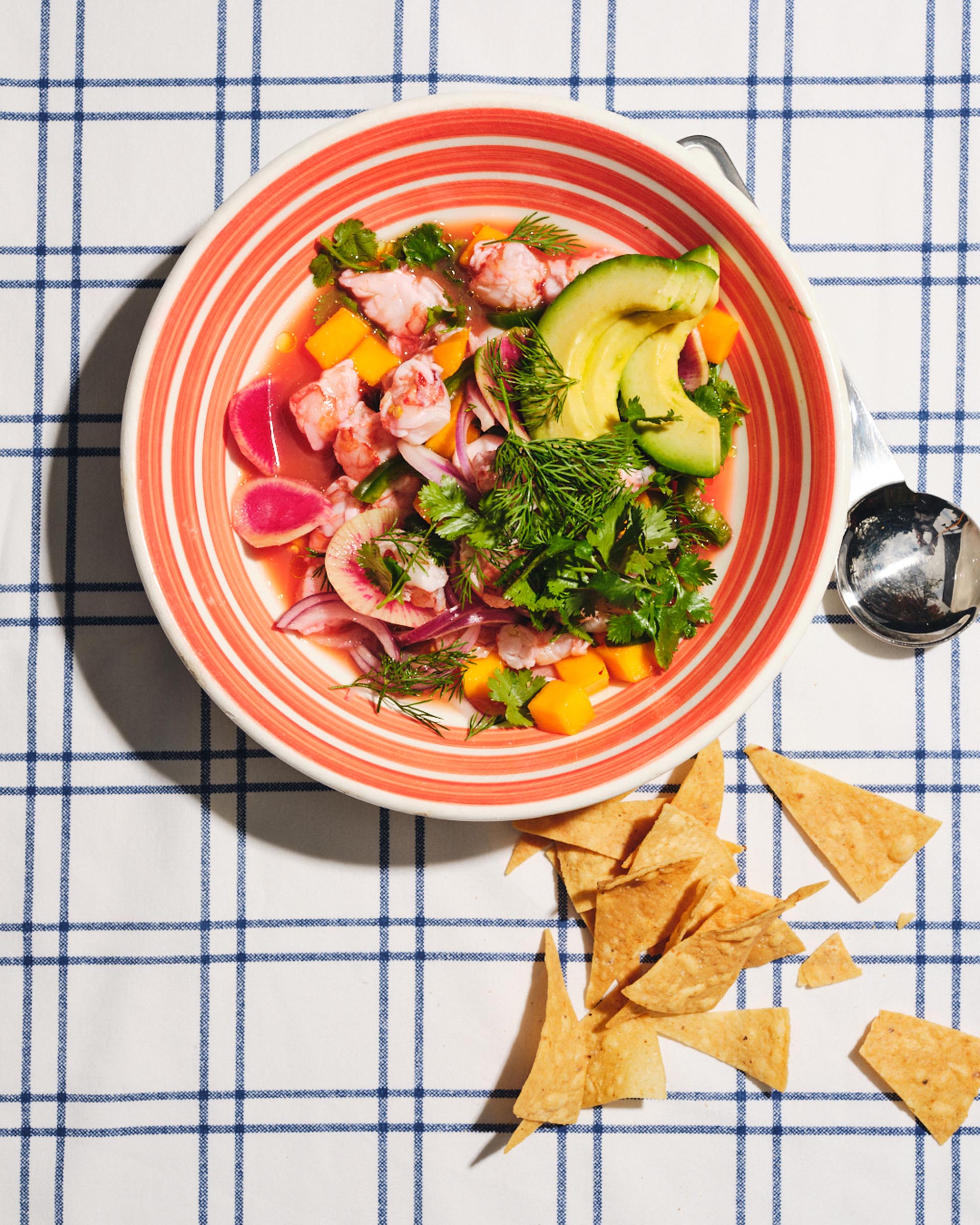 ceviche in pink bowl on checkered tablecloth with tortilla chips and spoon scattered next to it