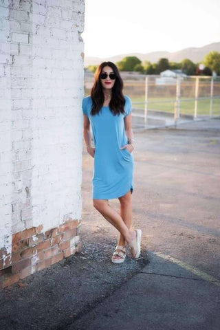 Female standing outside next to a white brick wall. She's wearing a kate blue dress from Brigitte Brianna and white sneakers.