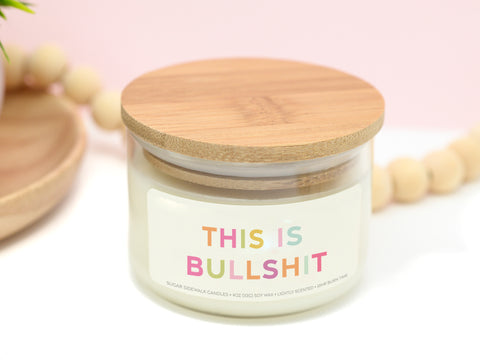 Candle in glass vessel with bamboo lid featuring label with colorful lettering that says this is bullshit