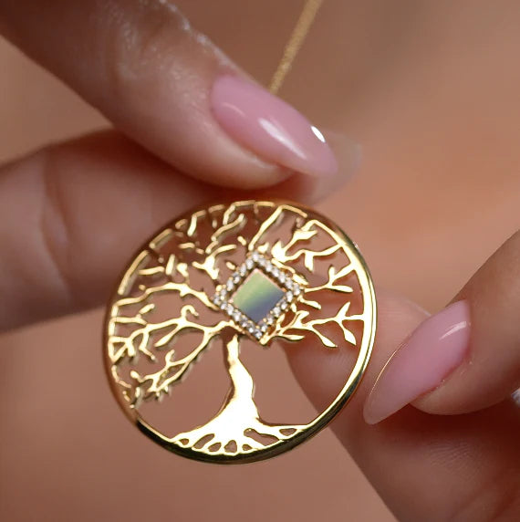 Gold tree of life pendant with an opal center, held by two fingers.