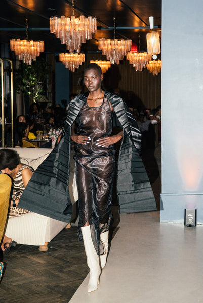 tall black runway model on runway wearing black cape under chandelier