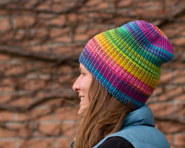 A profile shot of Catherine (that’s me!) wearing a brightly coloured, ribbed hat. The colorus are variegated and transition from blue at the brim to pink, yellow, green, blue, and purple at the crown.