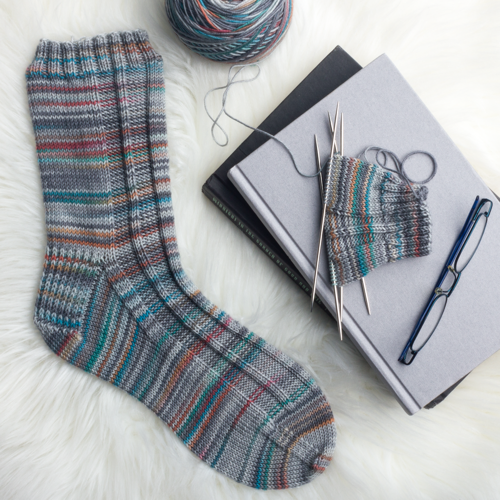 One handknit sock in variegated greys with little pops of orange, red, blue and green. The second sock is in progress, sitting beside a pair of reading glasses on a pile of books.