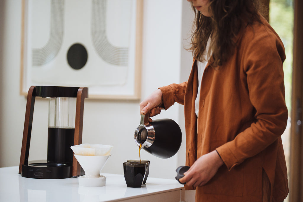 A woman pours coffee from a Ratio Eight thermal carafe into a glass.