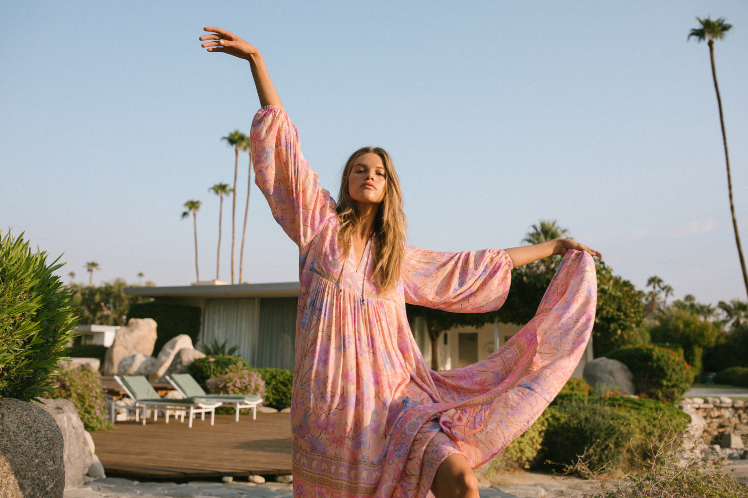 Spell Garden Party campaign model, Kim Riekenberg, posing on a wooden deck in a lush Palm Springs backyard while wearing pink pumps with the pale pink musk Spell Butterfly Boho Maxi Dress, featuring neckties and elasticated sleeve cuffs. 