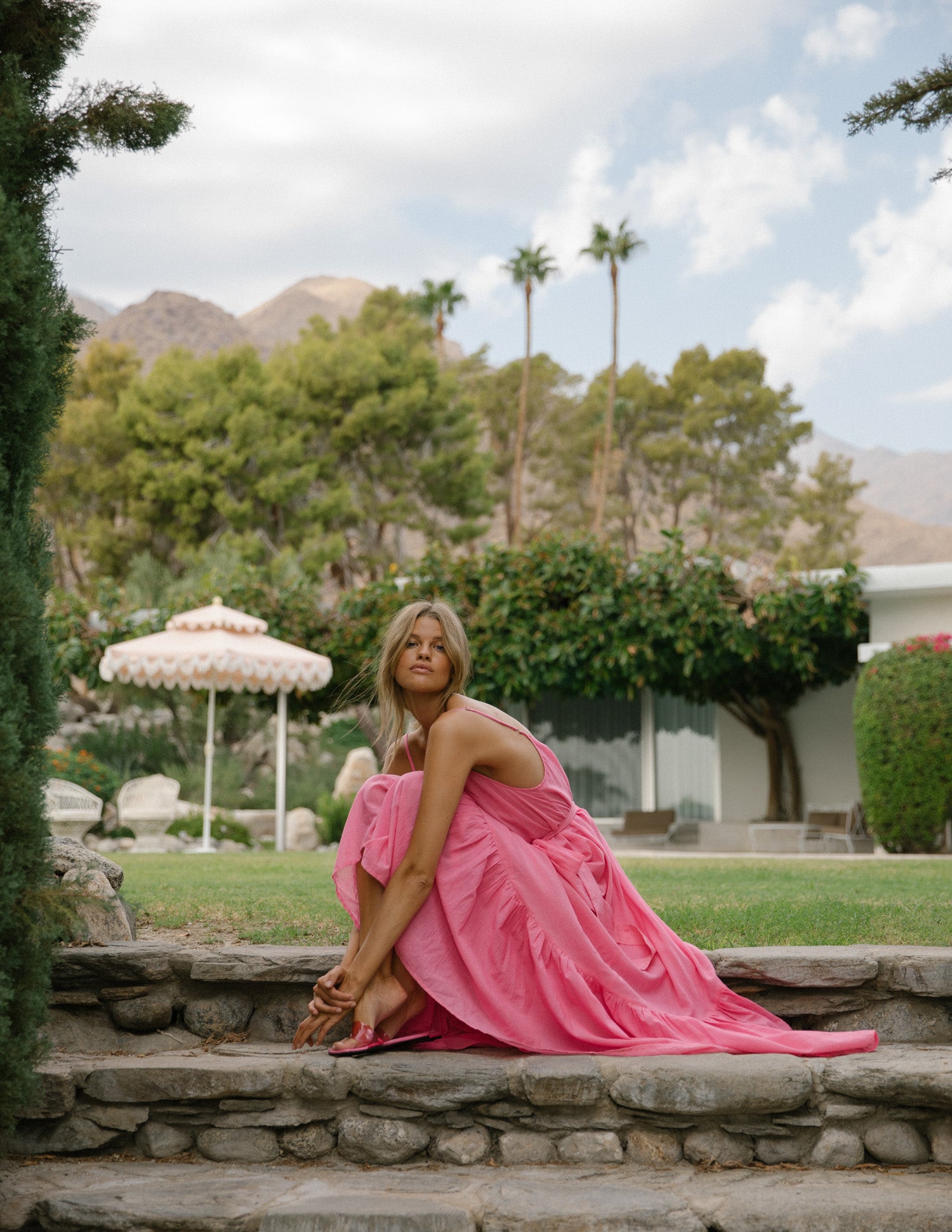 Spell Garden Party campaign model, Kim Riekenberg, frolicking through a lush Palm Springs backyard while wearing pink slides with the candy-coloured Spell Azalea Strappy Maxi Dress, featuring a detachable waist tie with belt loops and a deep v-neckline. 