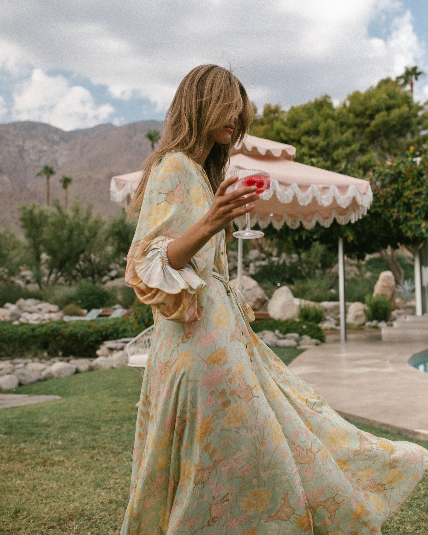 Spell Garden Party campaign model, Kim Riekenberg, dancing on lush green grass next to a Palm Springs pool with a cocktail in hand while wearing mint green slides and the mint green botanical Spell Butterfly Gown, featuring a brass button down front and raglan sleeves. 