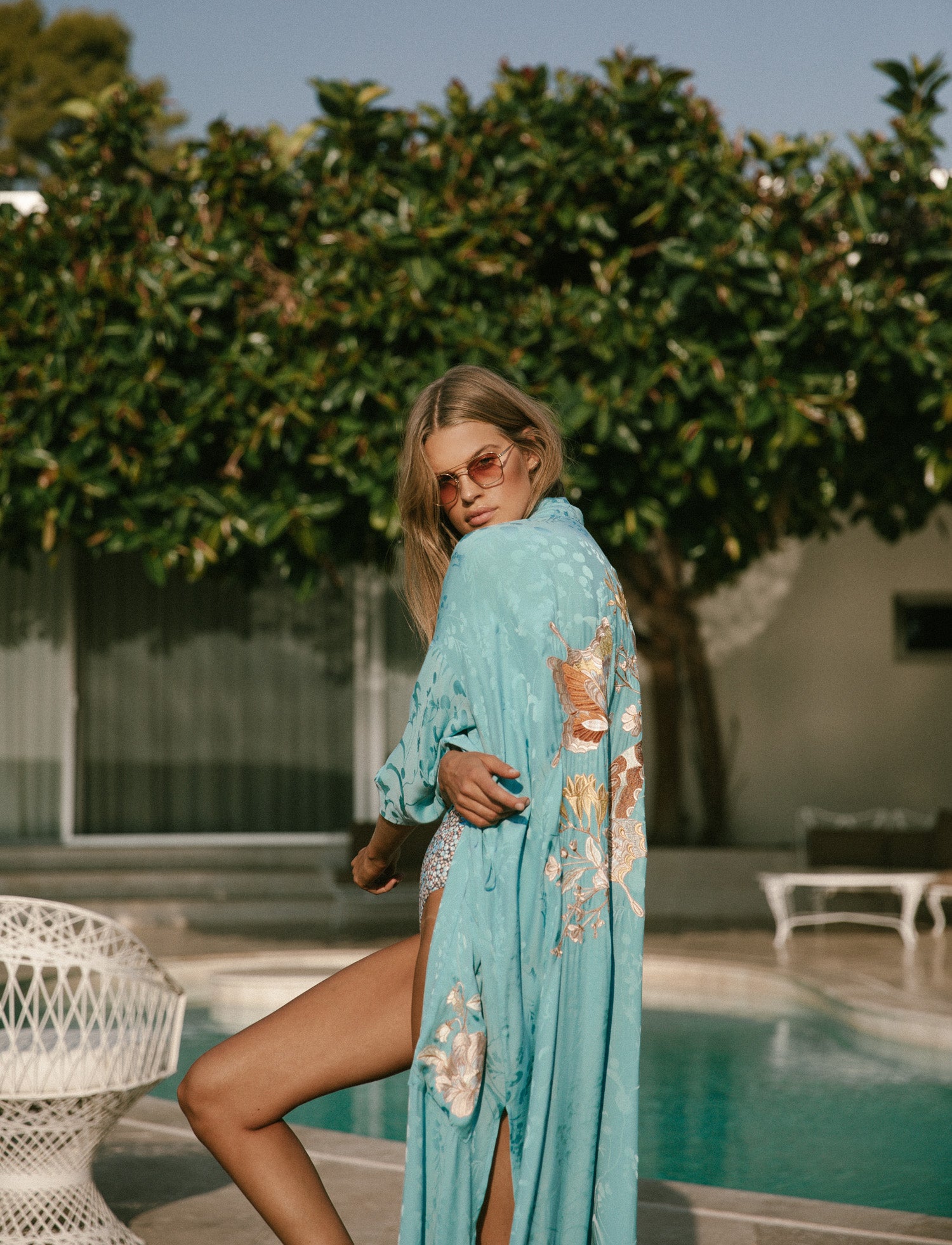 Spell Garden Party campaign model, Kim Riekenberg, sitting on a white steel chair on the outskirts of a pool in a leafy garden while wearing rose-tinted sunglasses, the Spell Poppy One Piece and the sapphire Spell Atrium Embroidered Maxi Robe, featuring an embroidery on the back and patch pockets. 