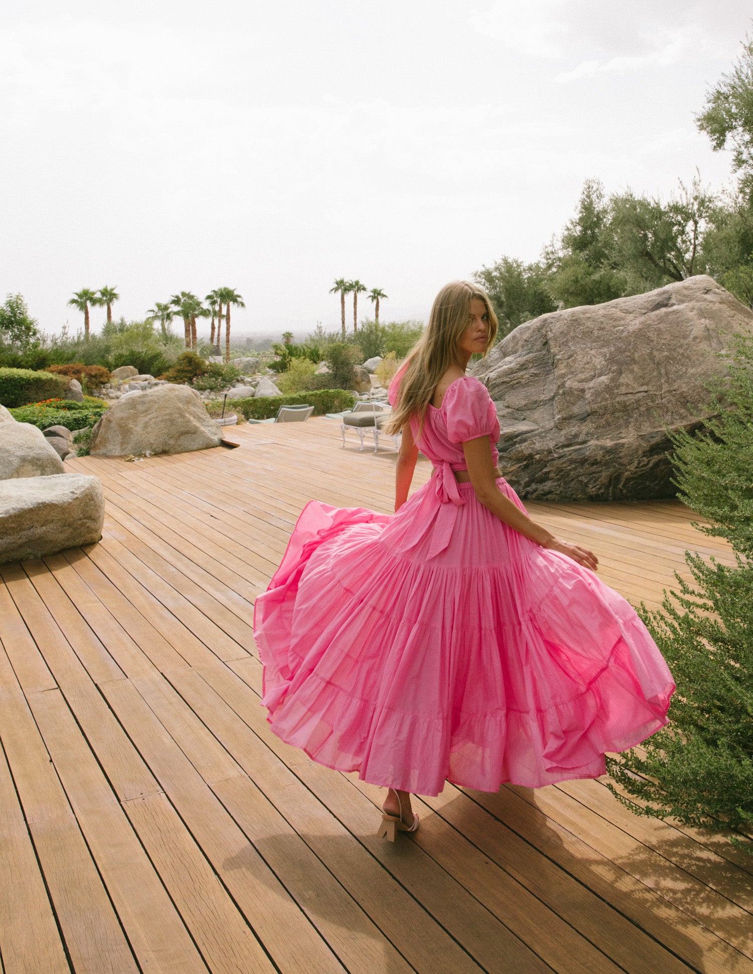 Spell Garden Party campaign model, Kim Riekenberg, walking on a wooden deck through a lush Palm Spiring front yard while wearing strappy white pumps with the candy-coloured Spell Azalea Cropped Blouse with the matching Azalea Tiered Skirt, featuring an elasticated waistband. 