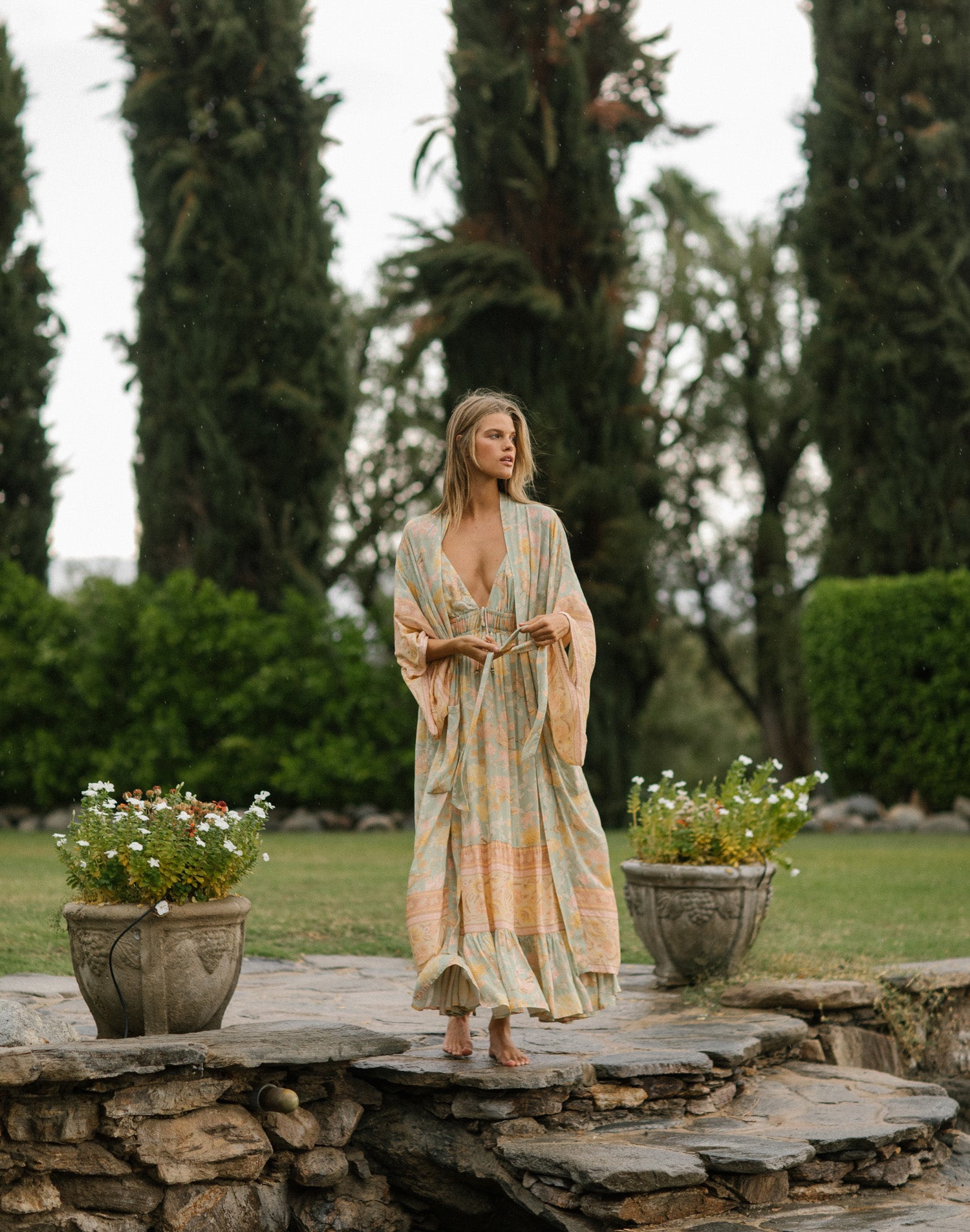 Spell Garden Party campaign model, Kim Riekenberg, walking on slated stones in a lush Palm Springs backyard while wearing the Spell Soiree Butterfly Dress with the matching mint green botanical Spell Butterfly Maxi Robe, featuring kimono-inspired sleeves and a detachable waist tie. 