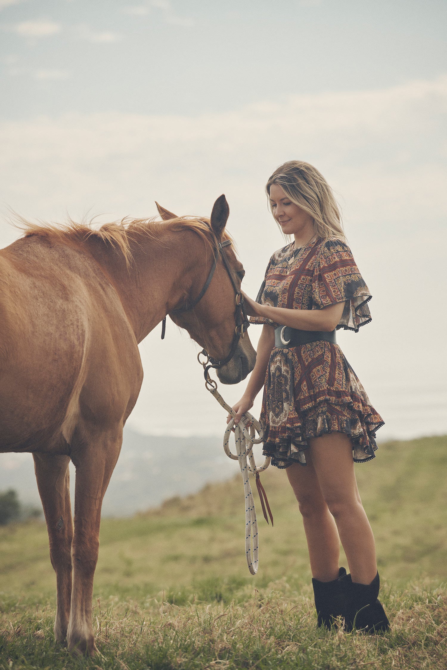 Jeweller and campaign model, Lucie Ferguson, posing in a green pasture with horses while wearing gold jewellery, black boots and the Spell regal Rumour Print Mini Dress with flutter sleeves and a gathered frill hem. 