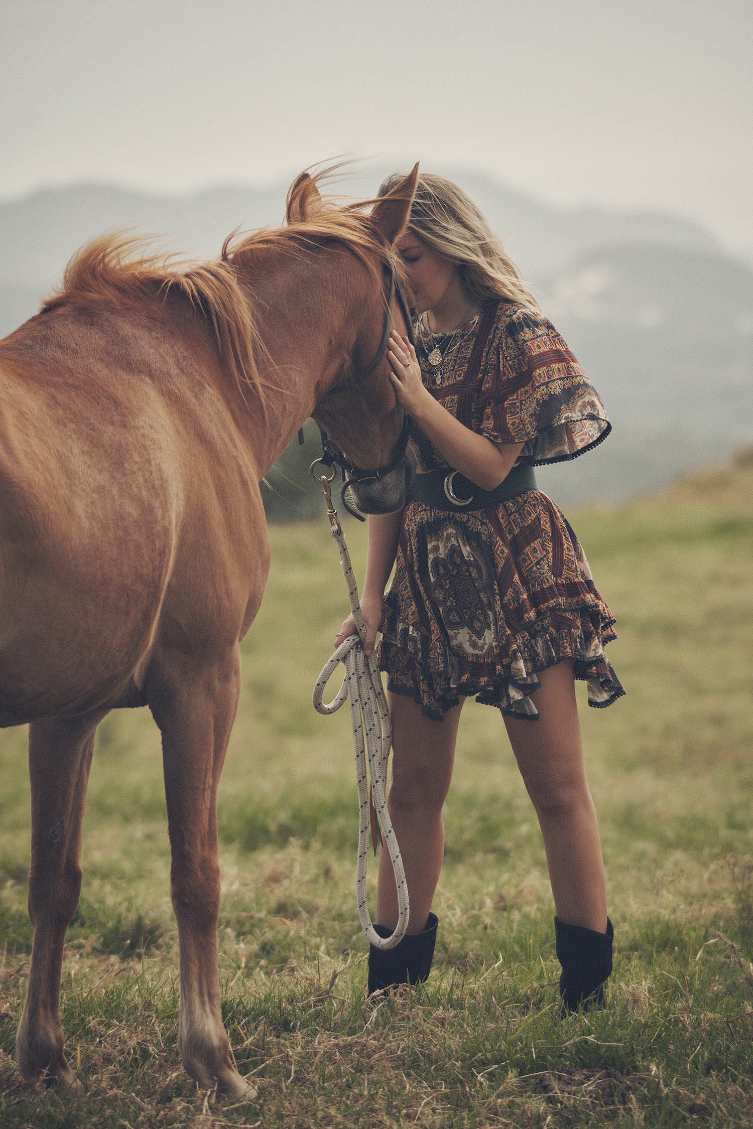 Jeweller and campaign model, Lucie Ferguson, posing in a green pasture with horses while wearing gold jewellery, black boots and the Spell regal Rumour Print Mini Dress with flutter sleeves and a gathered frill hem. 