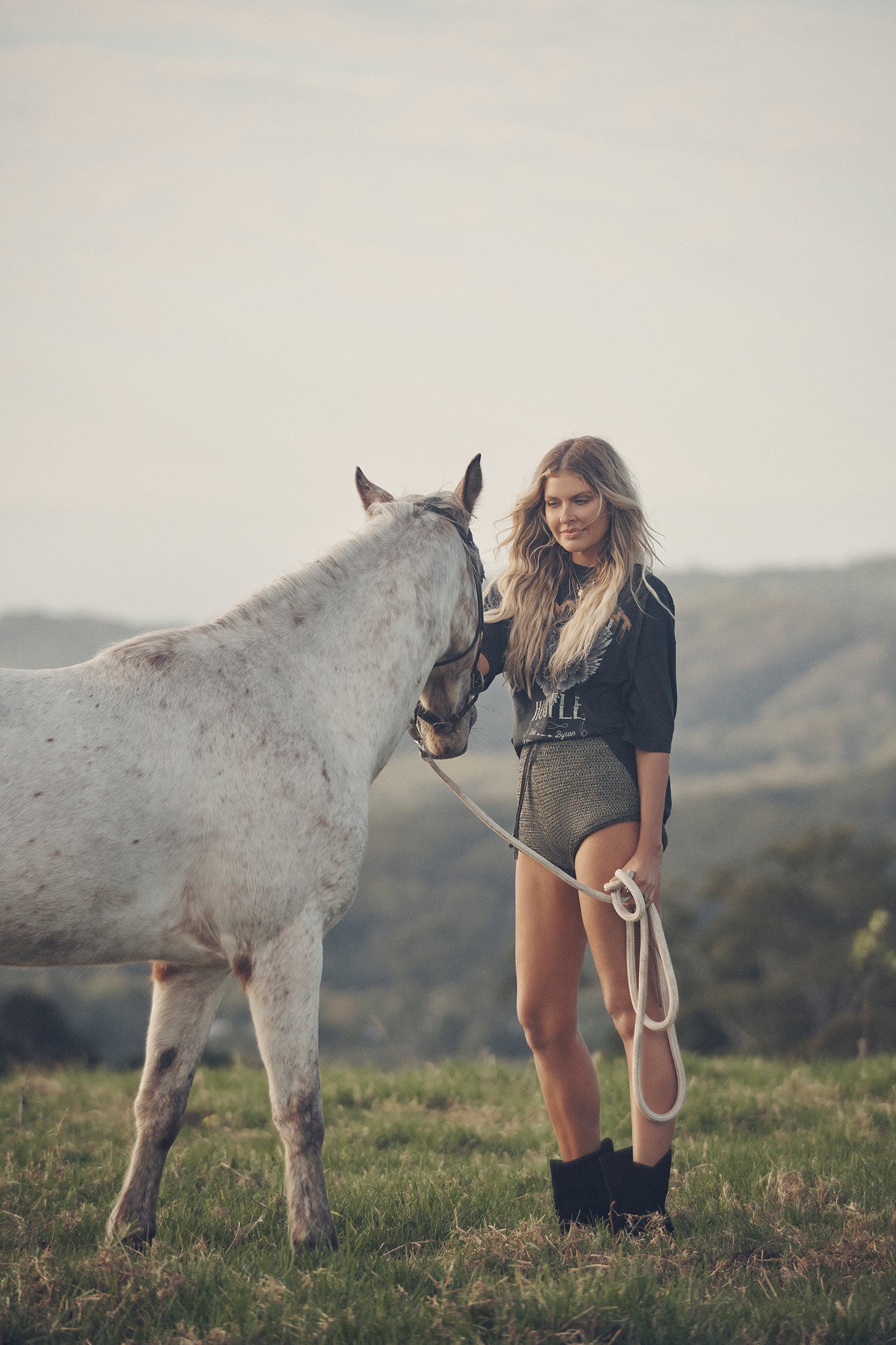 Influencer and model, Elle Ferguson, standing next to a white horse while wearing gold necklaces, the Spell Lucy Luna Earrings, oxyn Moonlight Shorts and the oversized biker charcoal Cult Tee Dress. 
