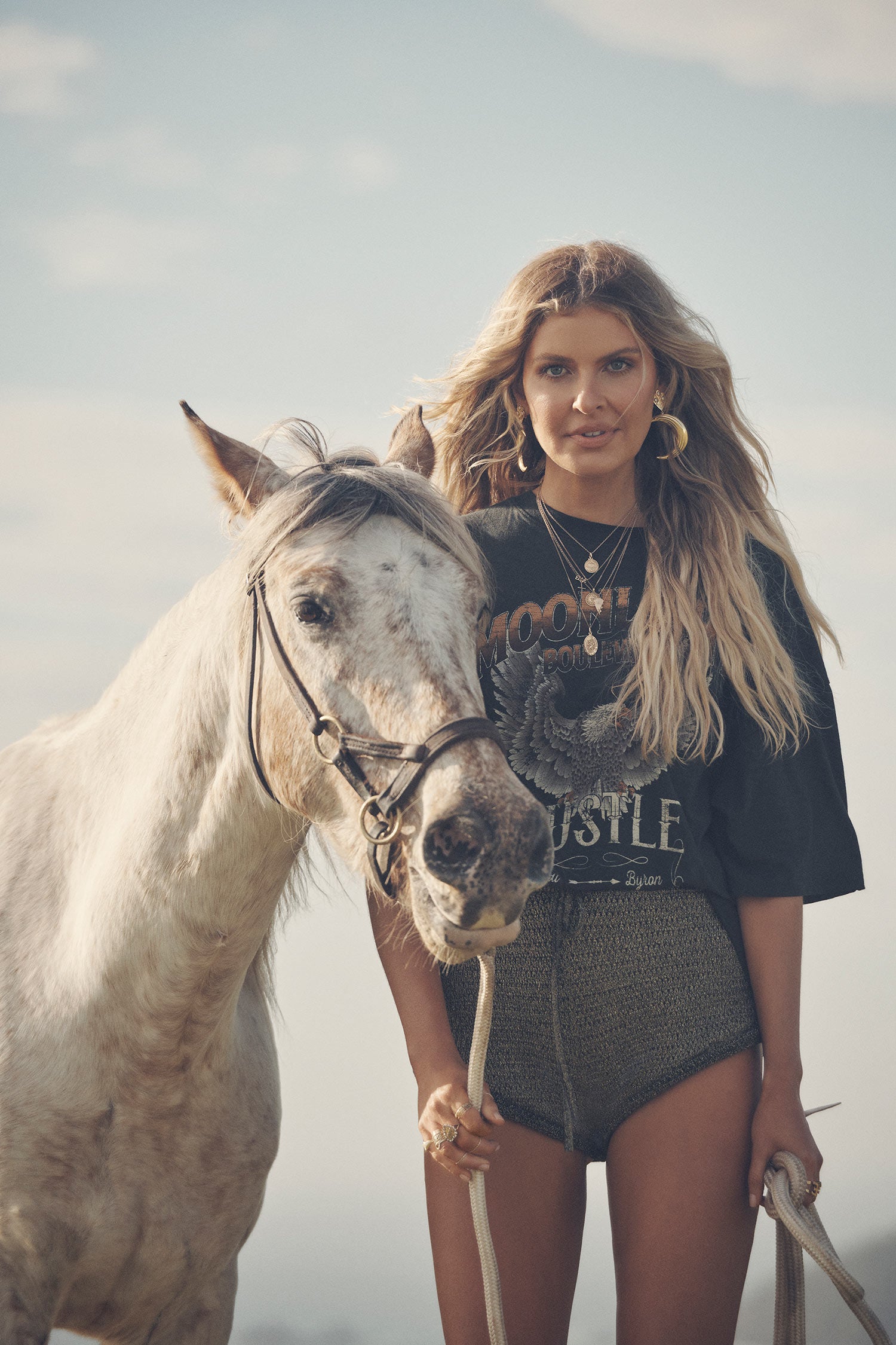 Influencer and model, Elle Ferguson, standing next to a white horse while wearing gold necklaces, the Spell Lucy Luna Earrings, oxyn Moonlight Shorts and the oversized biker charcoal Cult Tee Dress. 