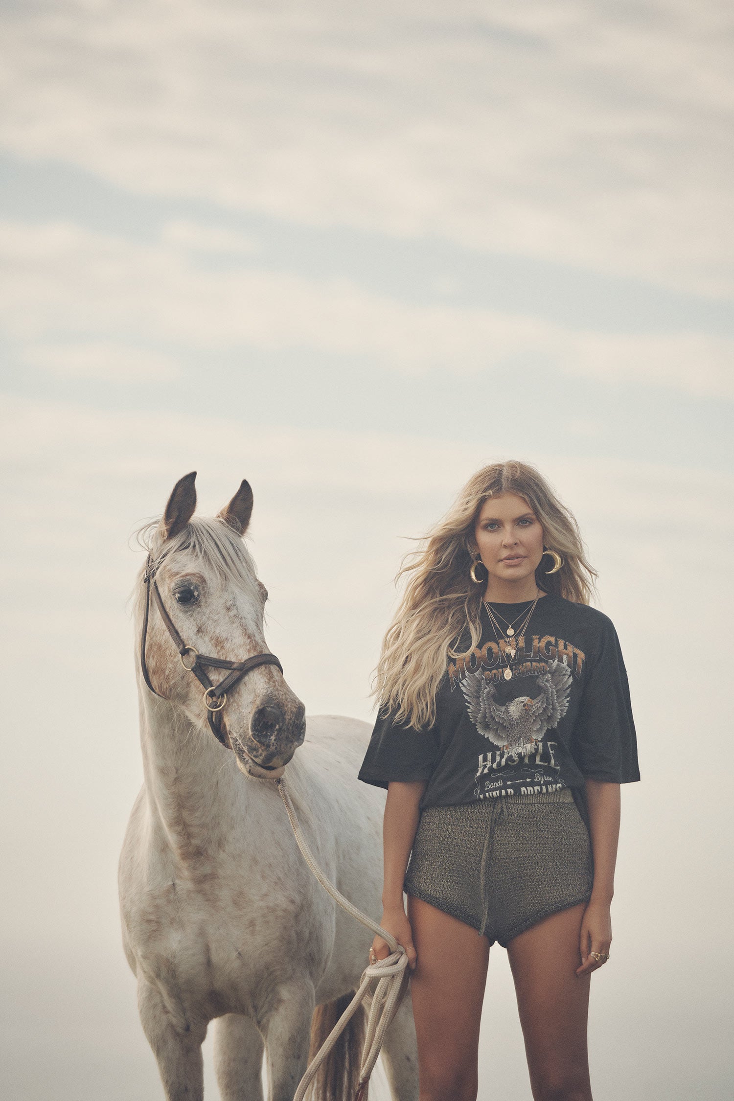 Influencer and model, Elle Ferguson, standing next to a white horse while wearing gold necklaces, the Spell Lucy Luna Earrings, oxyn Moonlight Shorts and the oversized biker charcoal Cult Tee Dress. 