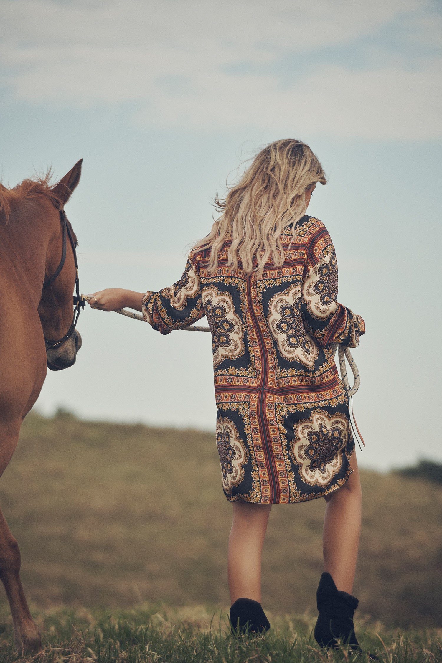 SpellJeweller and campaign model, Lucie Ferguson, posing in a green pasture with a brown horse while wearing gold jewellery, the Spell Cult Suede Slouch Boots and the Spell regal Rumour Print Shirt Dress.  