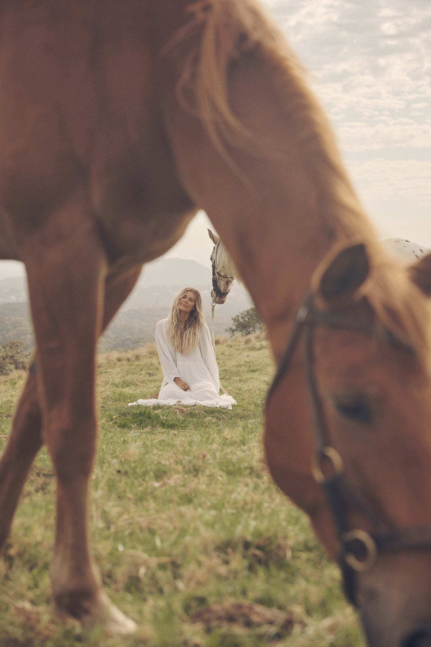 SpellInfluencer and model, Elle Ferguson, posing in a green pasture with a white horse while wearing gold jewellery, the Spell Cult Suede Slouch Boots with the white, tiered Moonflower Gown featuring frills and elasticated cuffs. 